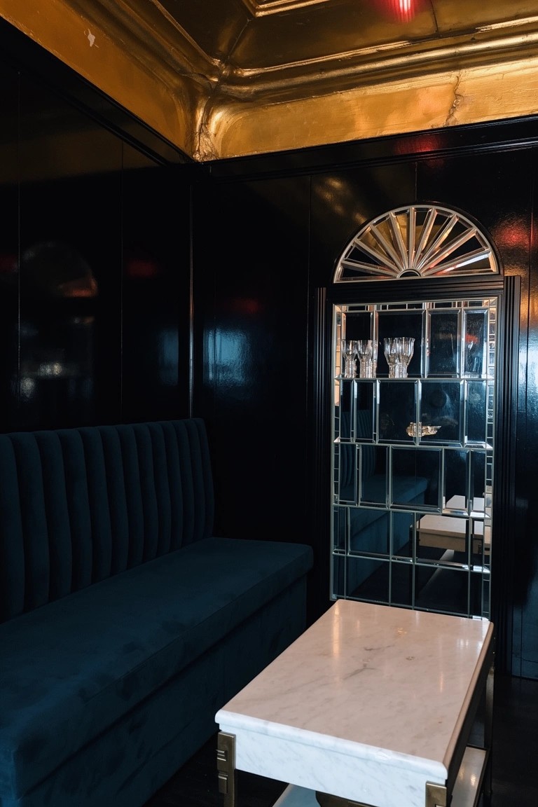 Black-paneled lounge nook with blue velvet bench, low marble table, and tall arched mirrored cabinet holding glassware