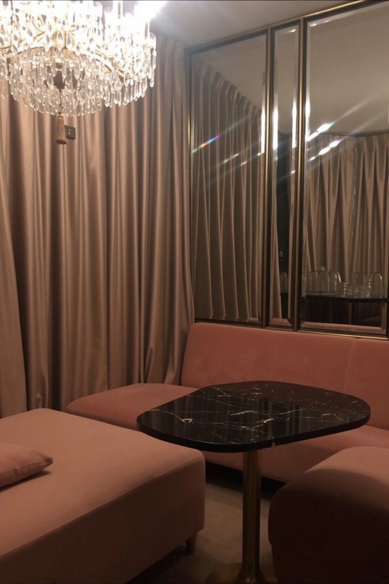 Pink velvet low sofas and ottomans around a black marble table in a curtained room with mirrors and chandelier
