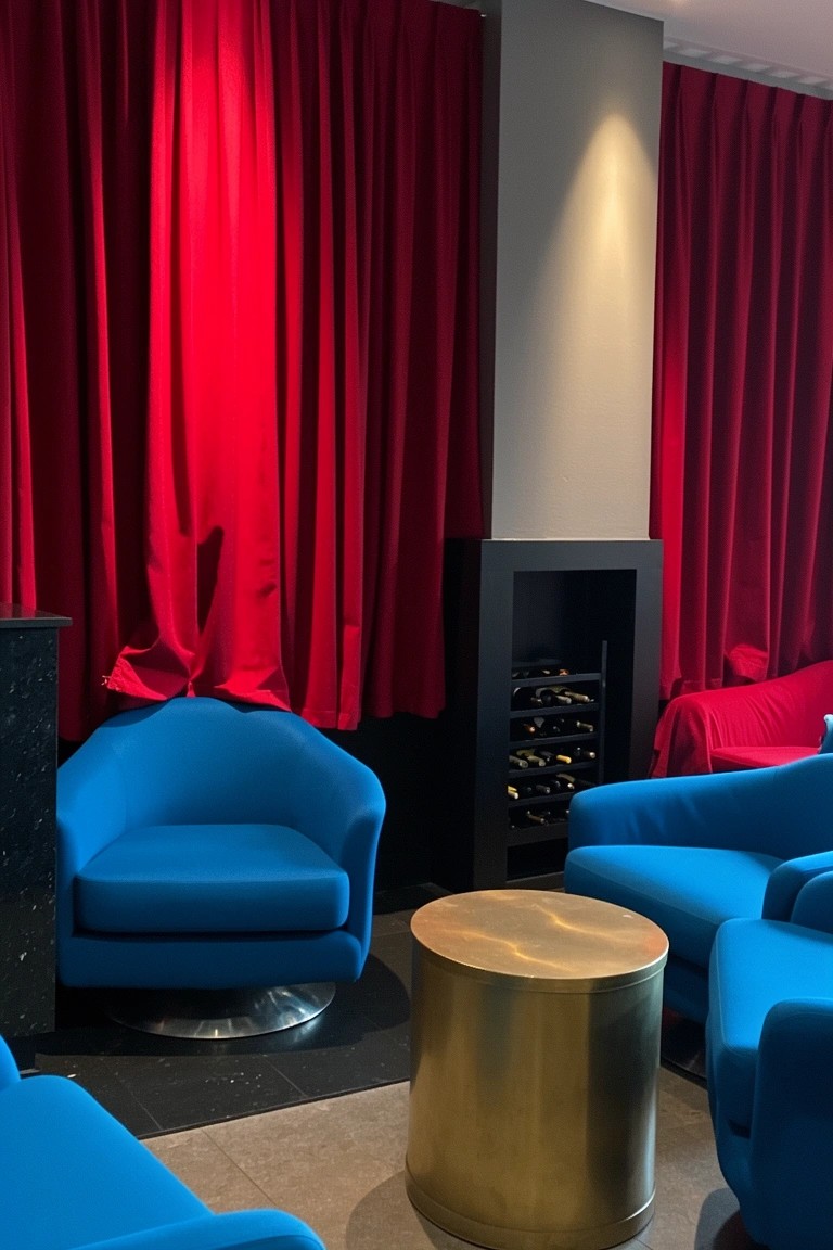 Intimate lounge with red velvet curtains framing blue swivel chairs around a brass drum table and bar cabinet