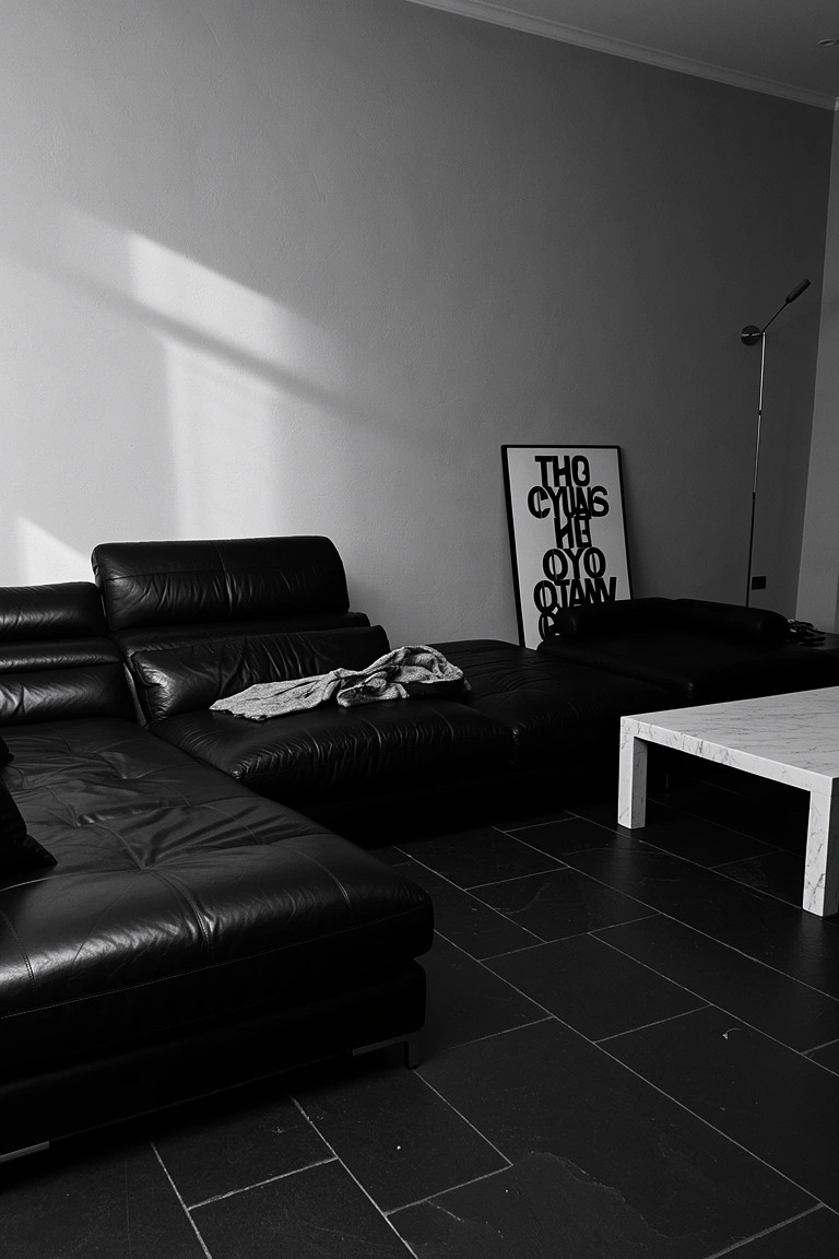Black and white photo of a minimalist sitting room with a large black leather L-shaped sofa, white marble coffee table, graphic wall poster, and tiled floor