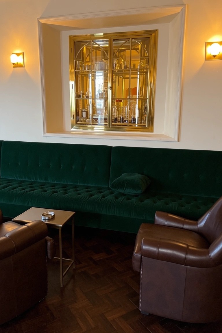 Ornate gold-framed glass cabinet displaying bottles above green velvet sofa and leather chairs in a lounge area