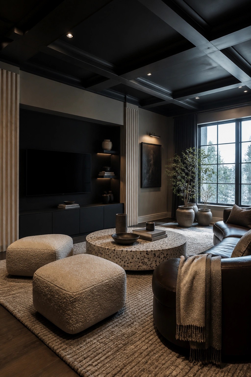 A living room with black walls and ceiling, a built-in media console with TV, two beige textured ottomans, a round terrazzo coffee table, and a brown leather sofa on a large woven rug.
