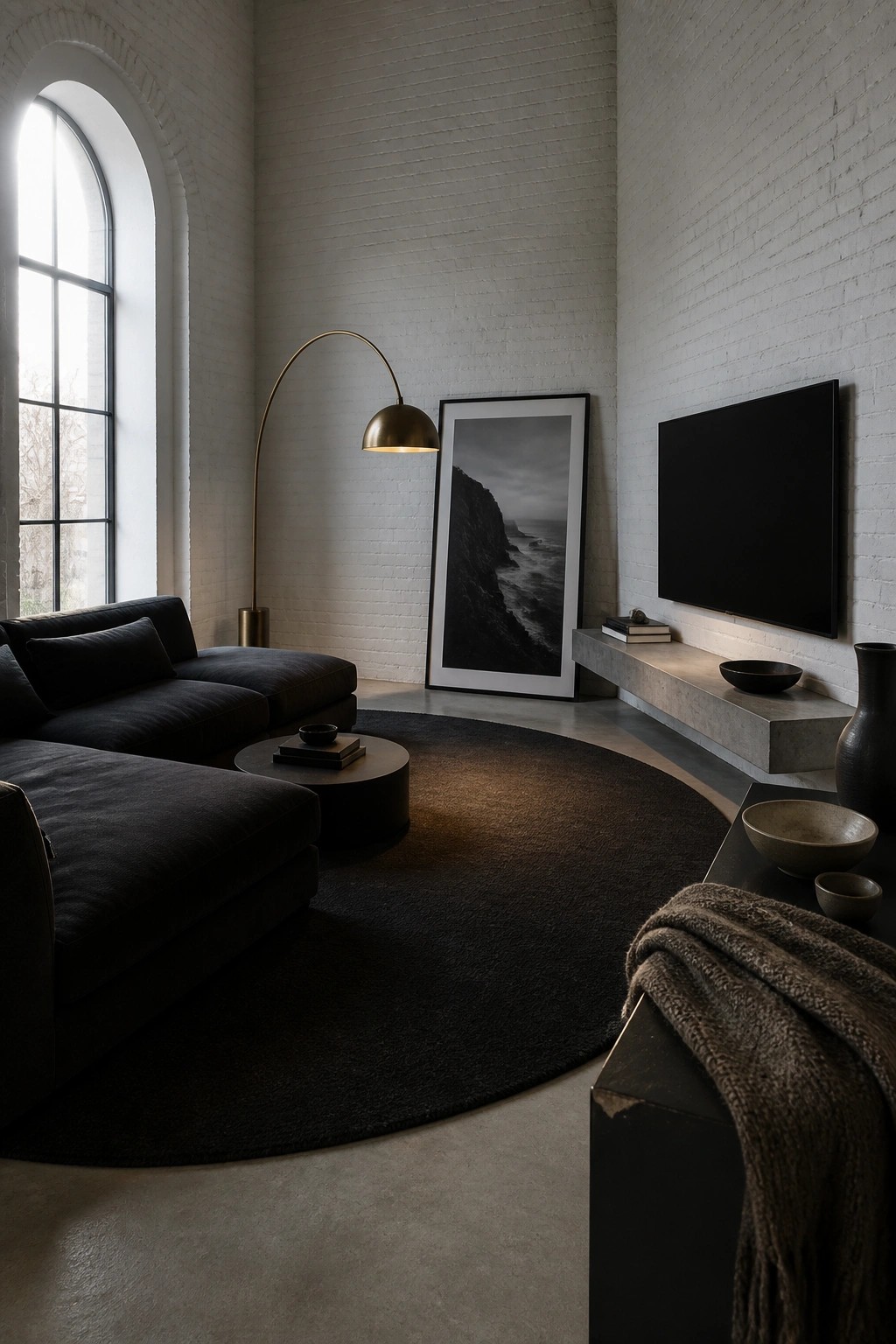 A living room with a dark sectional sofa on a large round black rug, white brick walls, an arched window, and a wall-mounted television above a floating concrete console.