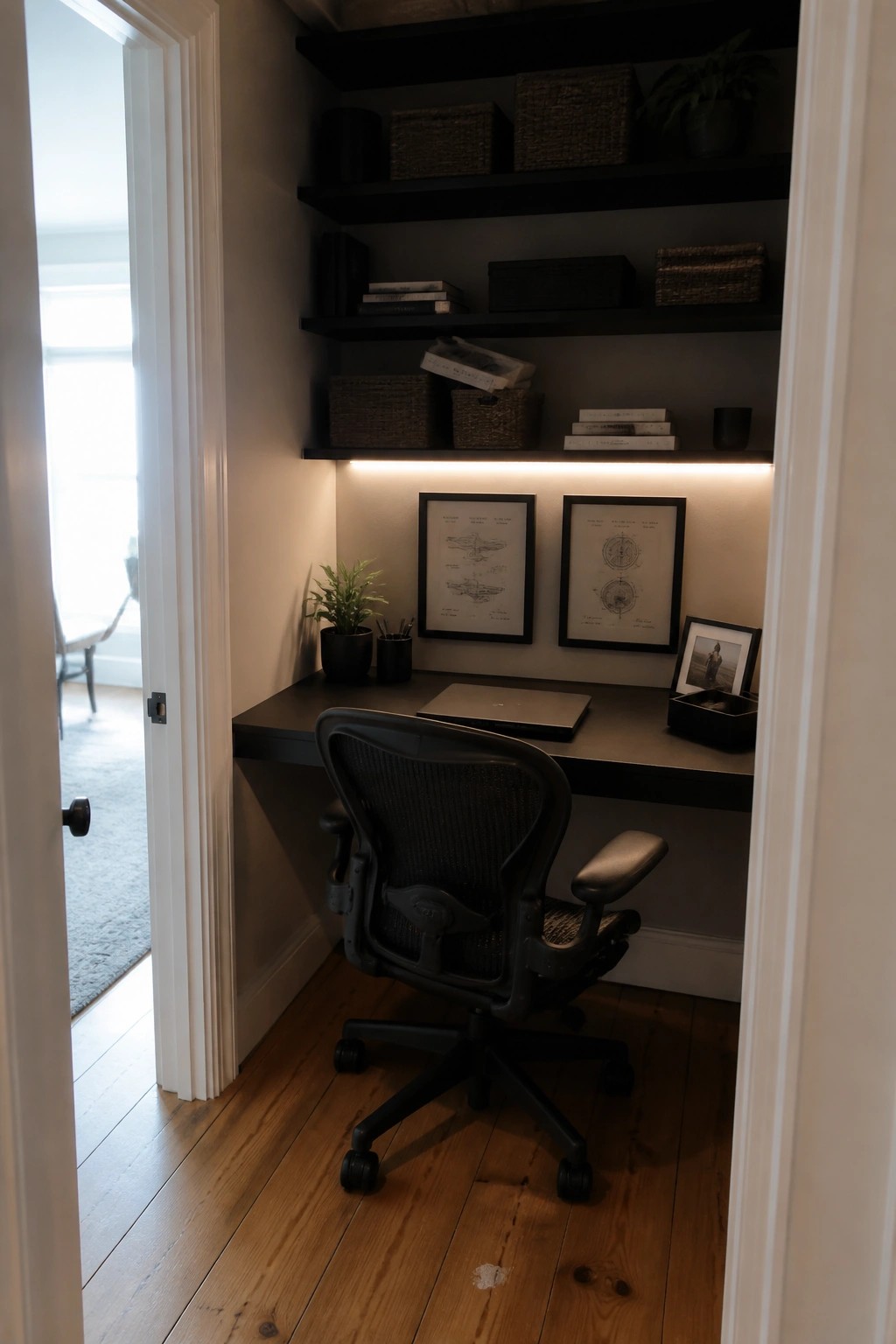 A narrow home office nook inside a closet with black floating shelves above a matching desk, under-shelf lighting, framed prints, and a black ergonomic chair on wood flooring.