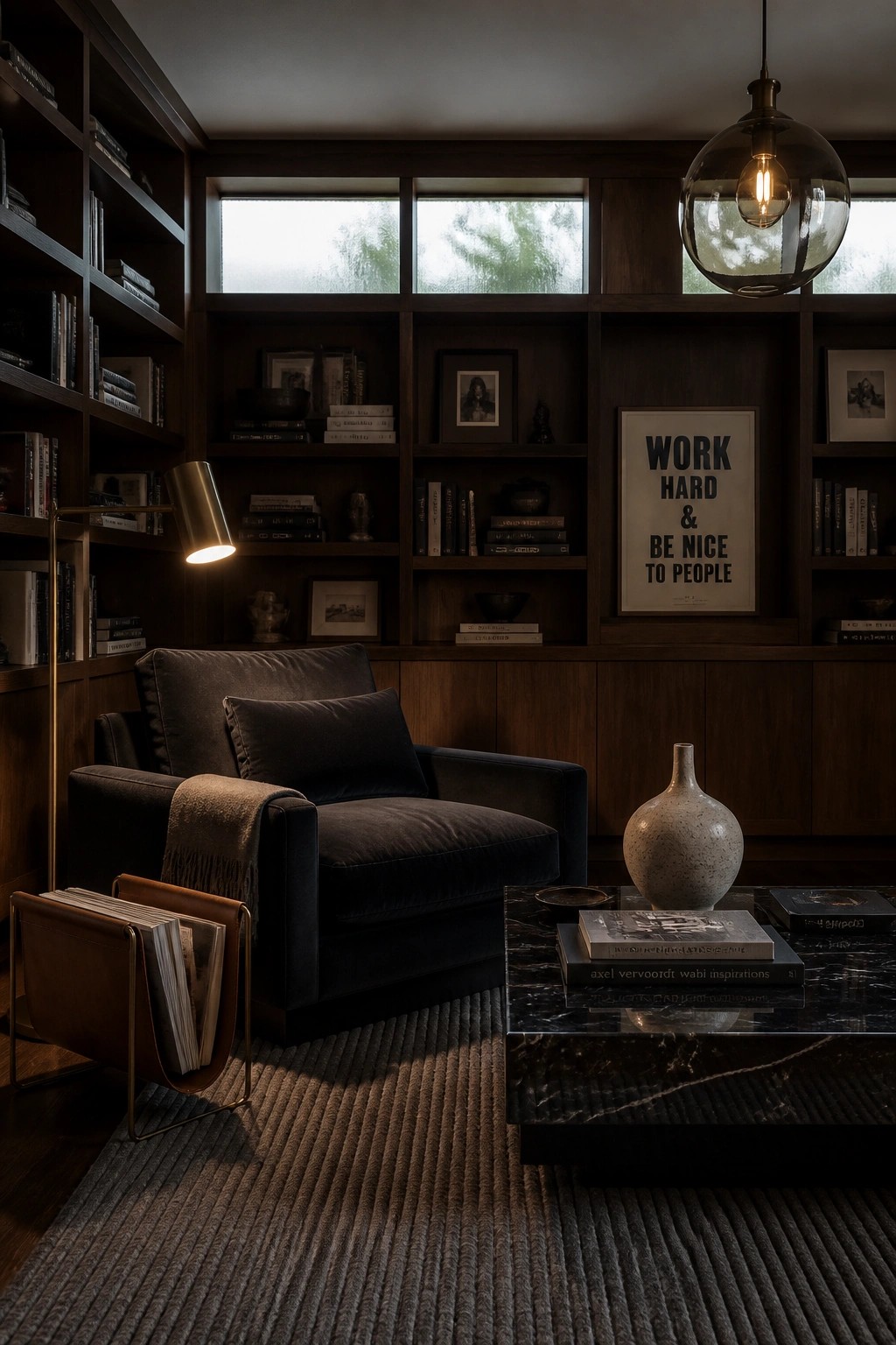 A dark living room with built-in wood bookshelves, a dark velvet armchair, a marble coffee table, and a large pendant light.