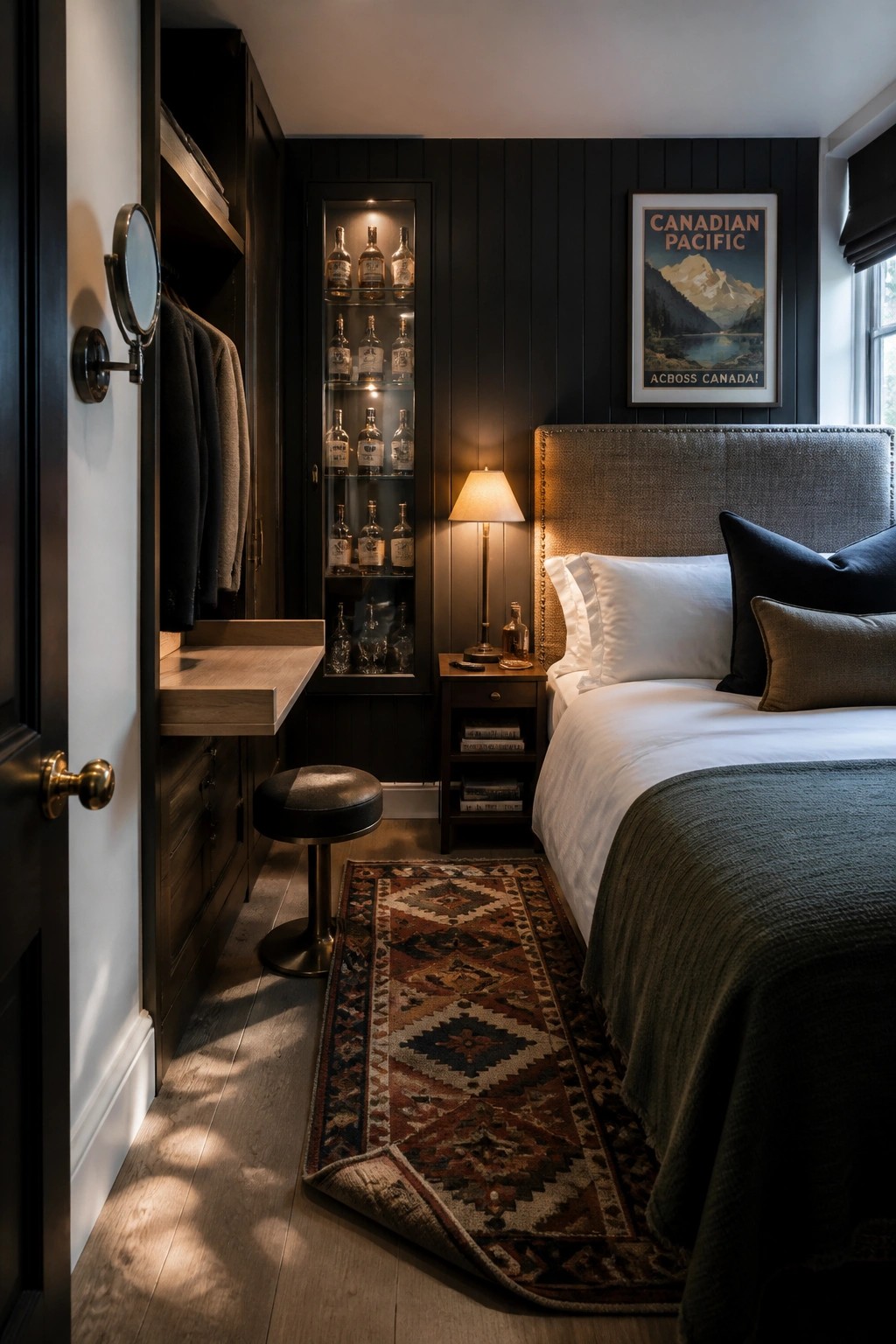 A compact bedroom with dark walls shows a bed with white bedding, a lit glass cabinet displaying bottles, built-in cabinetry with a fold-down desk, a stool, and a patterned rug on the wood floor.