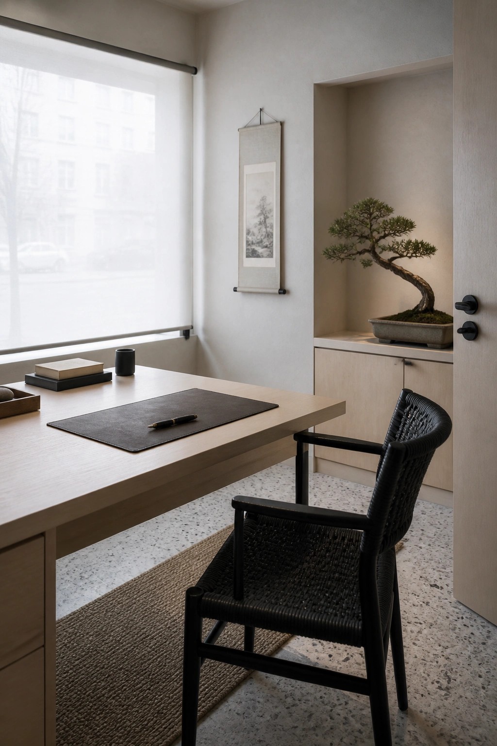 A small home office with a light wood desk, black woven chair, roller shade window, and a recessed niche holding a bonsai tree.