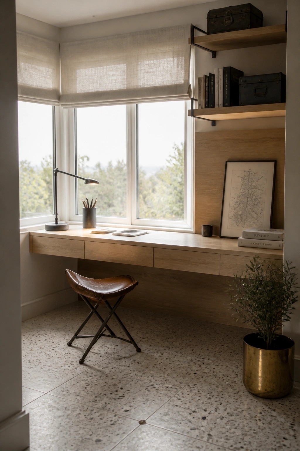A light wood built-in desk spans a large window wall in a home office, with a brown leather stool underneath and floating shelves holding books and boxes to the right.