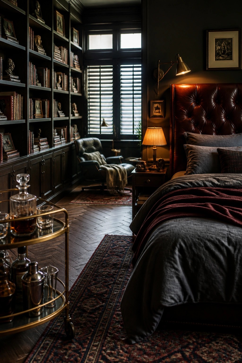 A dimly lit bedroom with dark walls, floor-to-ceiling built-in bookshelves filled with books, a large bed with a tufted leather headboard, and a bar cart in the foreground.