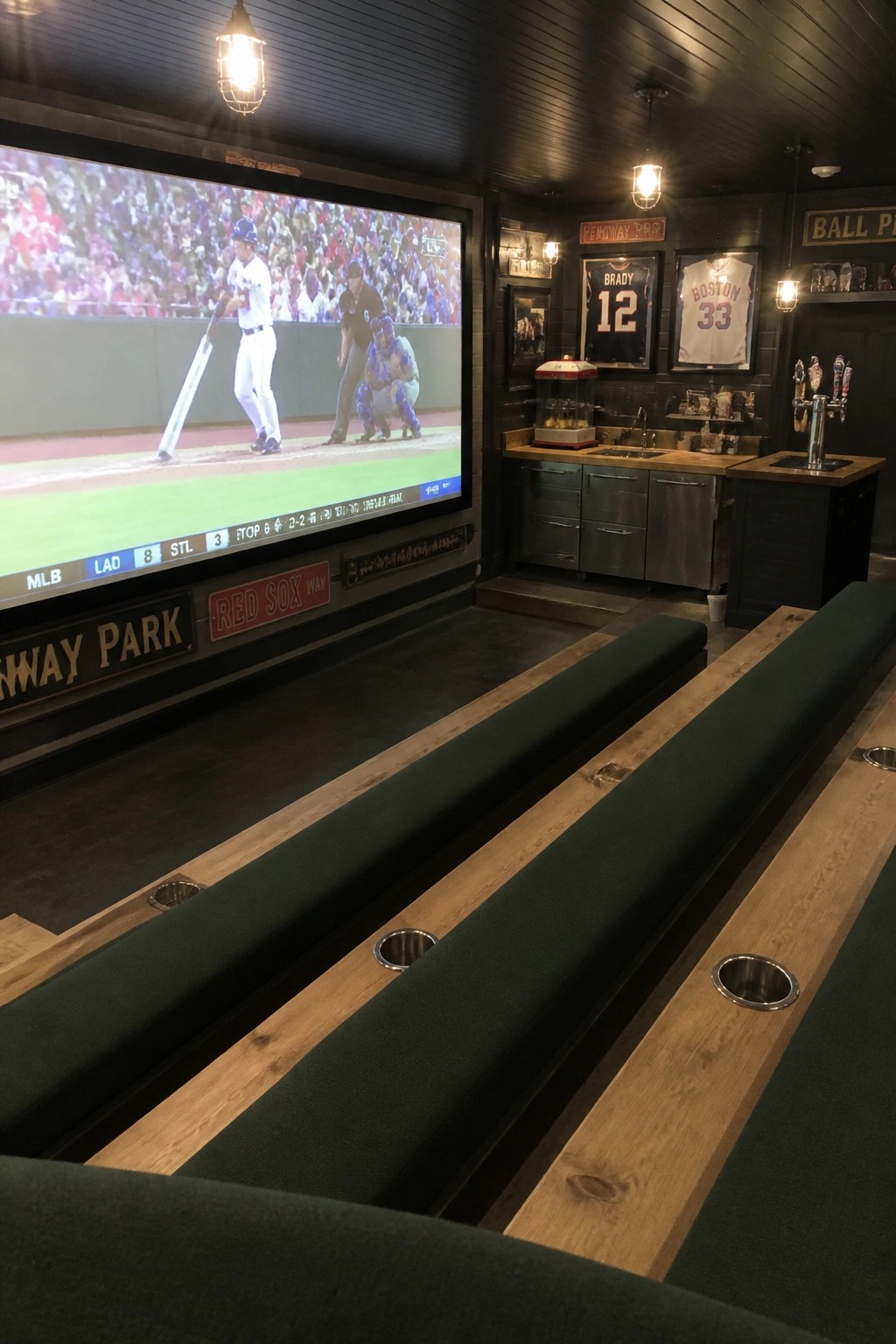 An indoor home theater room with a large projector screen showing a baseball game, tiered green upholstered benches with cup holders, and a bar area with sports jerseys on the wall.