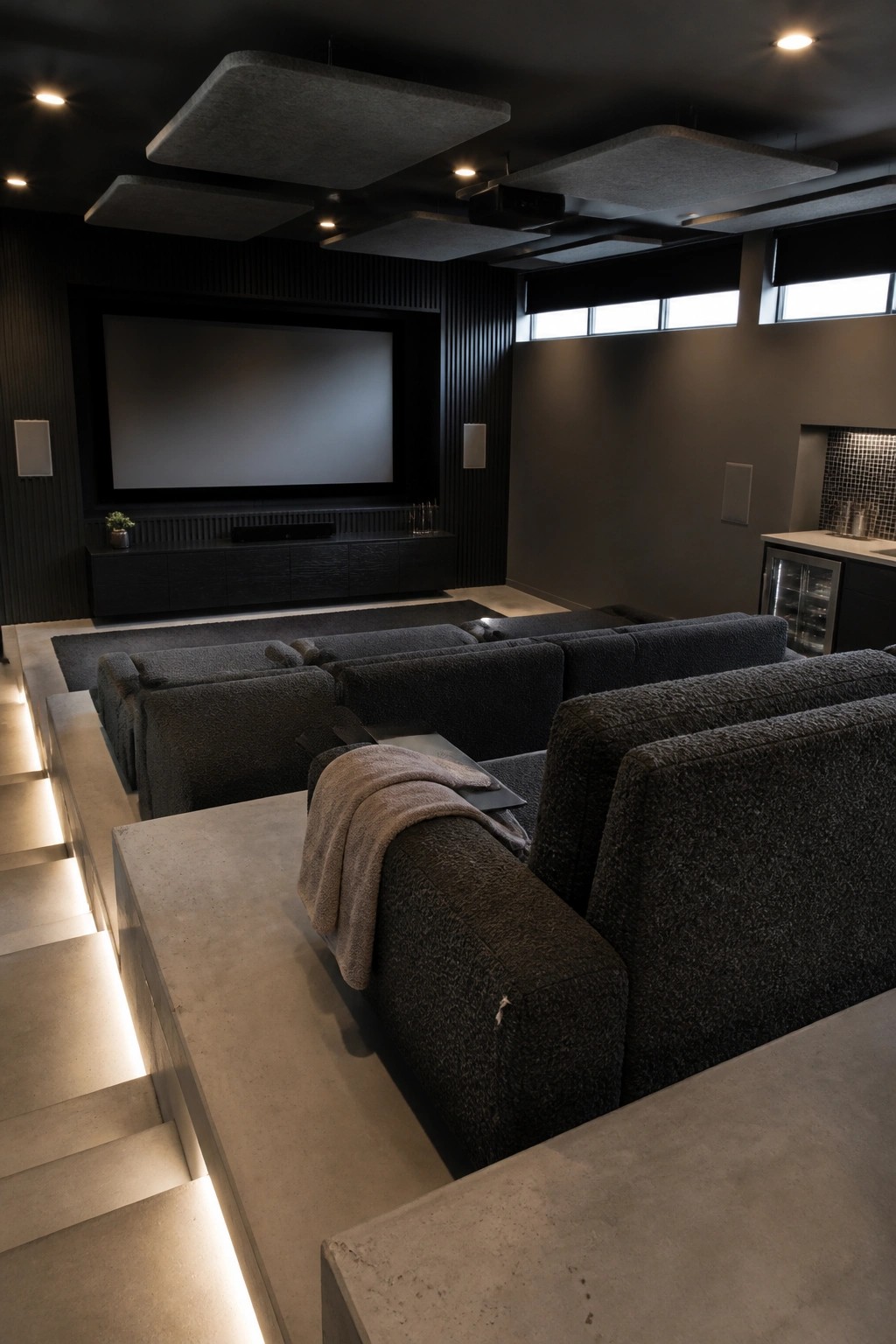 A home theater interior with tiered dark sectional seating on illuminated concrete steps facing a large projector screen, with acoustic panels on the ceiling.