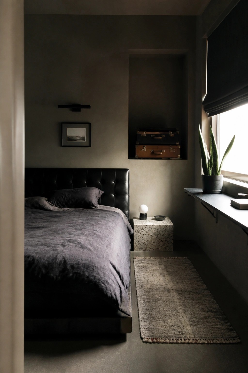 A dark minimalist bedroom featuring a black leather bed, a recessed wall niche with stacked suitcases, and a window ledge with a potted plant.