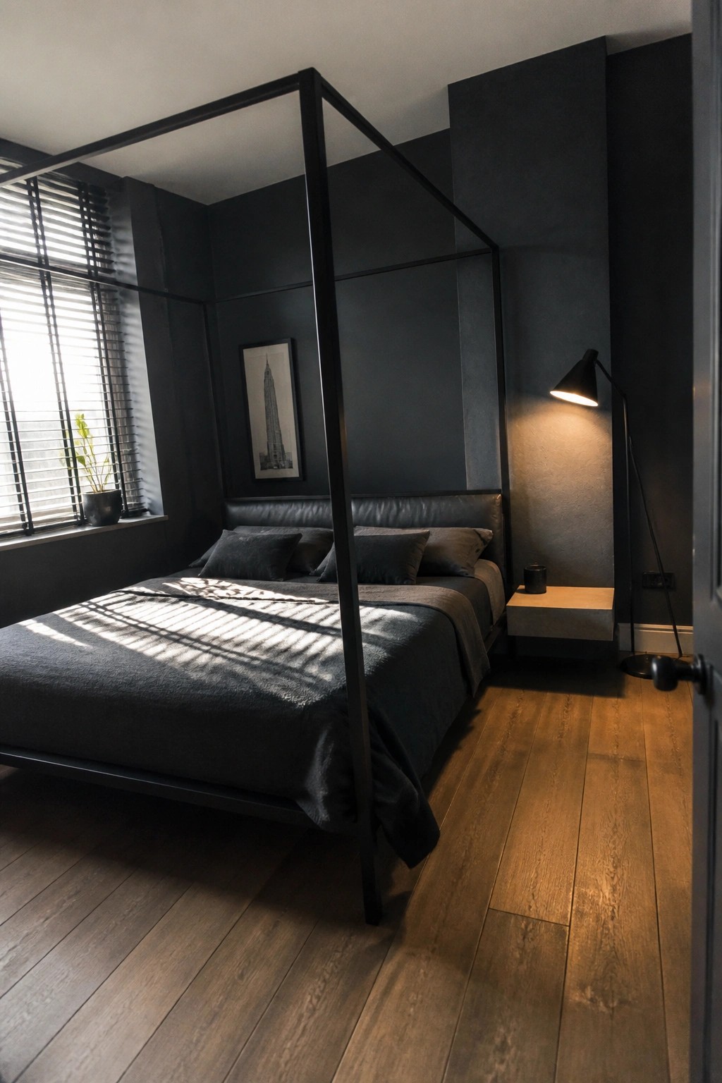 A minimalist bedroom with a black four-poster bed, dark bedding, dark walls, and light wood flooring.