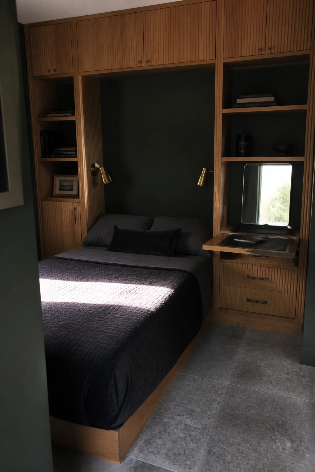 A small bedroom features a bed recessed into dark green walls and surrounded by light wood built-in cabinets, open shelves, and a fold-down desk with mirror on the right side.