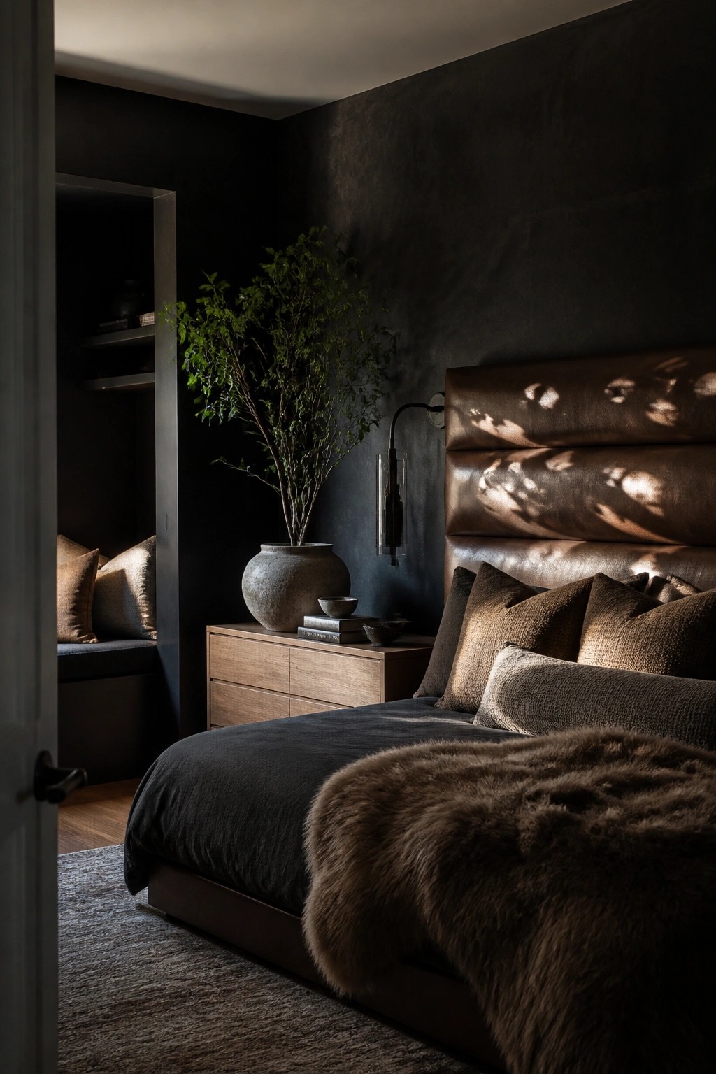 A dark bedroom with black walls, a brown leather headboard, layered pillows, a fur throw on the bed, a wooden nightstand, and a large potted plant.