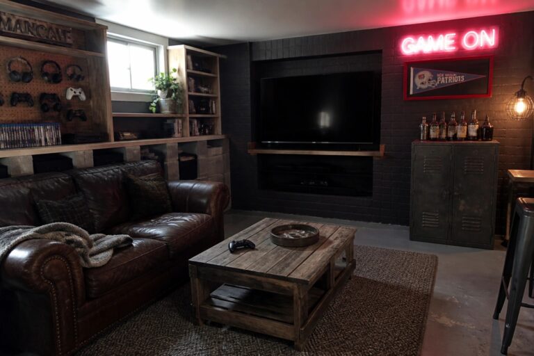 19 Budget Man Cave Ideas Built From DIY Shelving and Secondhand Furniture A man cave interior with a wall-mounted TV above a brick fireplace, wooden crate shelving filled with items, a metal locker, a brown leather sofa, and a rustic coffee table on a woven rug.