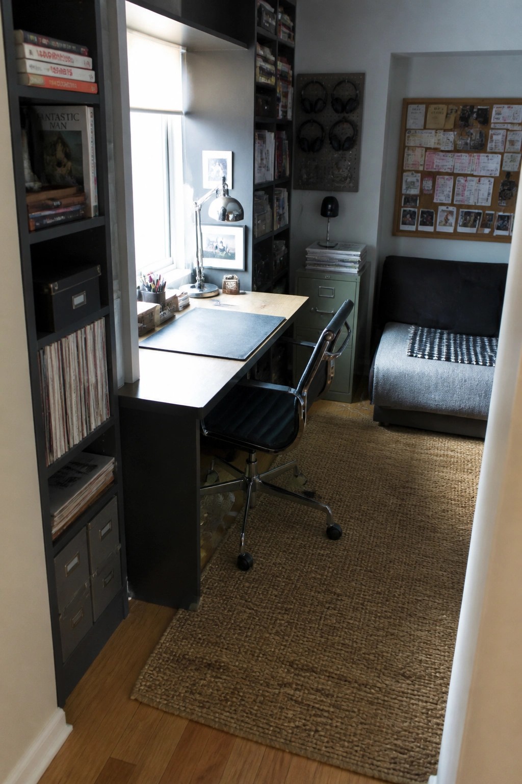 Compact home office with desk, black chair, bookshelves, daybed, corkboard, and woven rug.