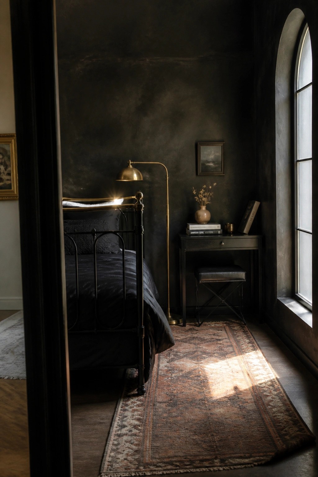 A dark bedroom featuring black walls, black bedding on a metal bed frame, a brass floor lamp, a small dark desk, and a patterned rug on the floor.