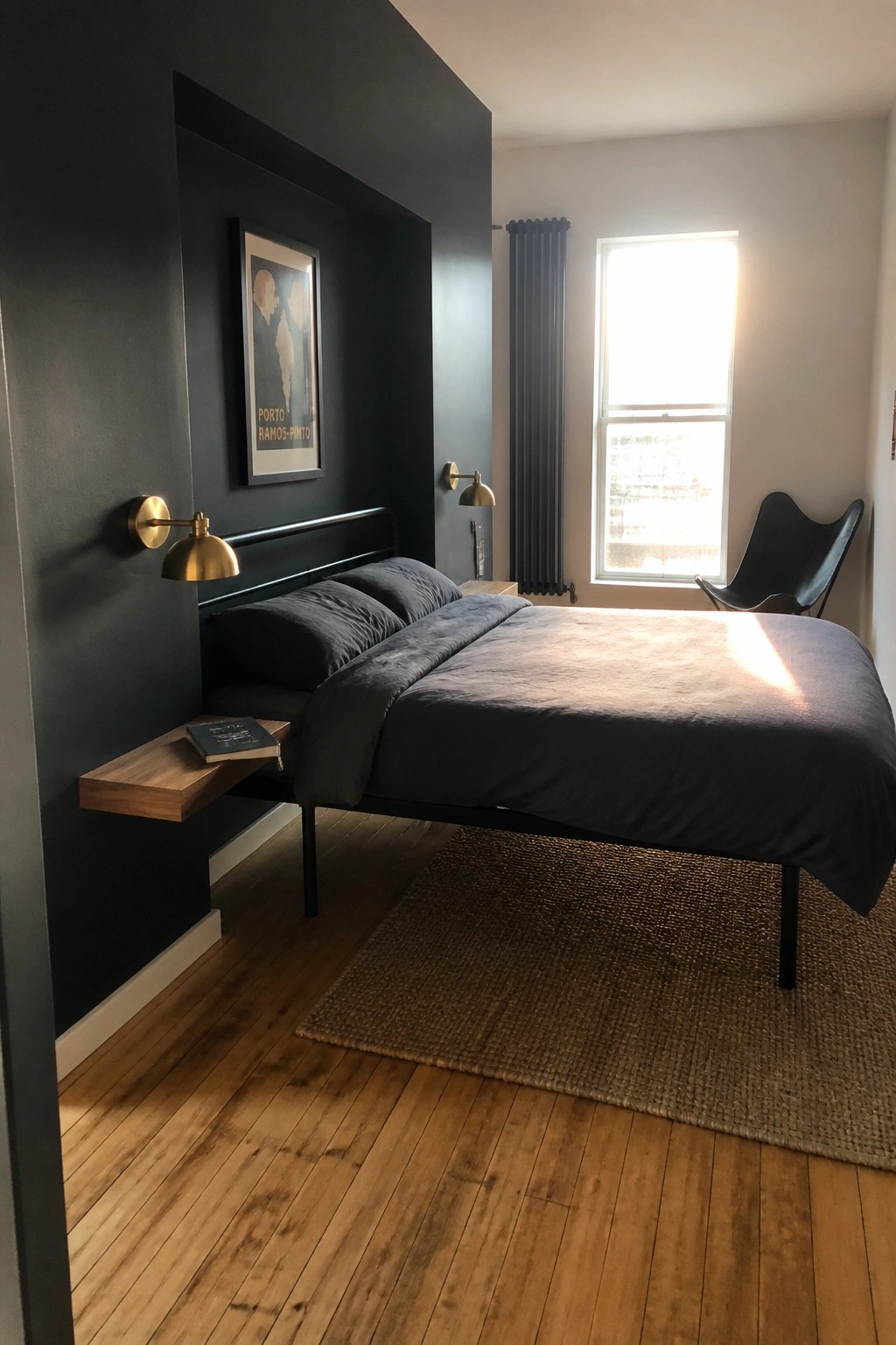 A bedroom with black walls, a bed with dark gray bedding, two brass wall sconces, and a floating wooden shelf beside the bed.