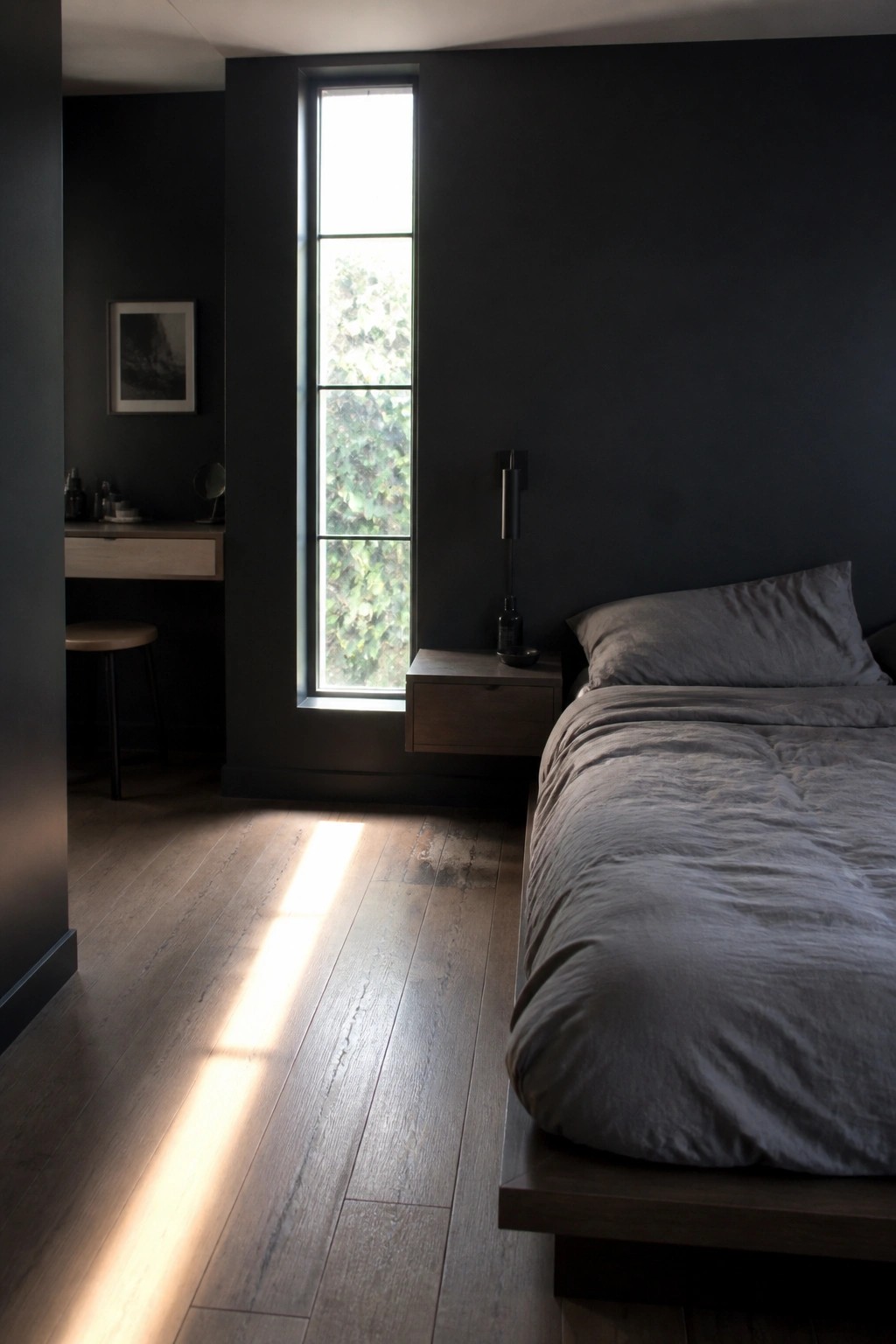 A bedroom with matte black walls, a tall narrow window, light brown wood flooring, a low platform bed with gray bedding, and a floating wood nightstand.
