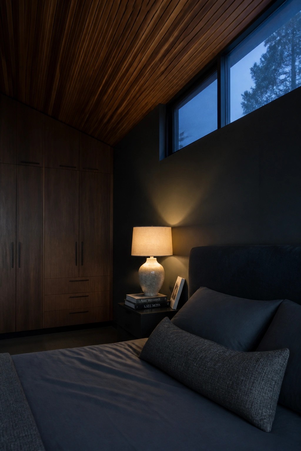 Bedroom with black walls, slatted wood ceiling, tall dark wood built-in cabinets, bed with dark gray bedding, and a lit table lamp on a nightstand.