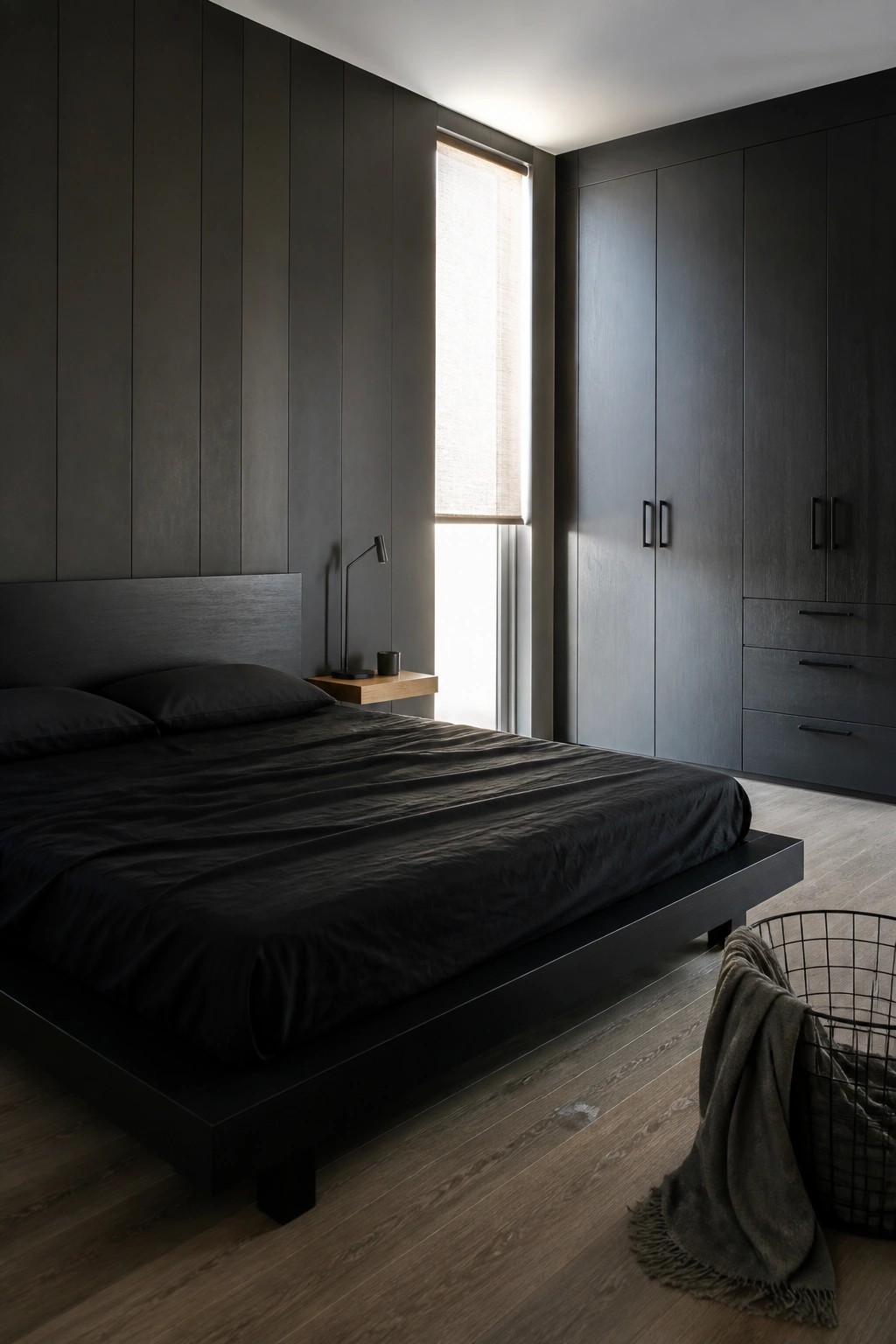 A bedroom with a low black platform bed, dark walls, a built-in wardrobe, and a window with a roller shade.