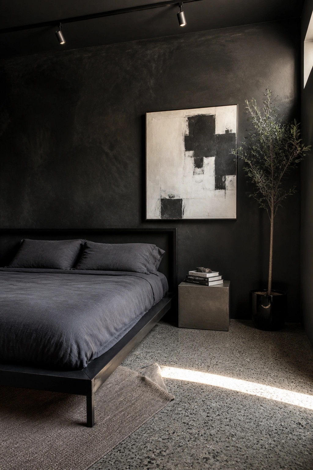 A bedroom featuring a low black platform bed with dark gray bedding against textured black walls, an abstract black and white painting, a concrete side table with books, and a tall potted tree.