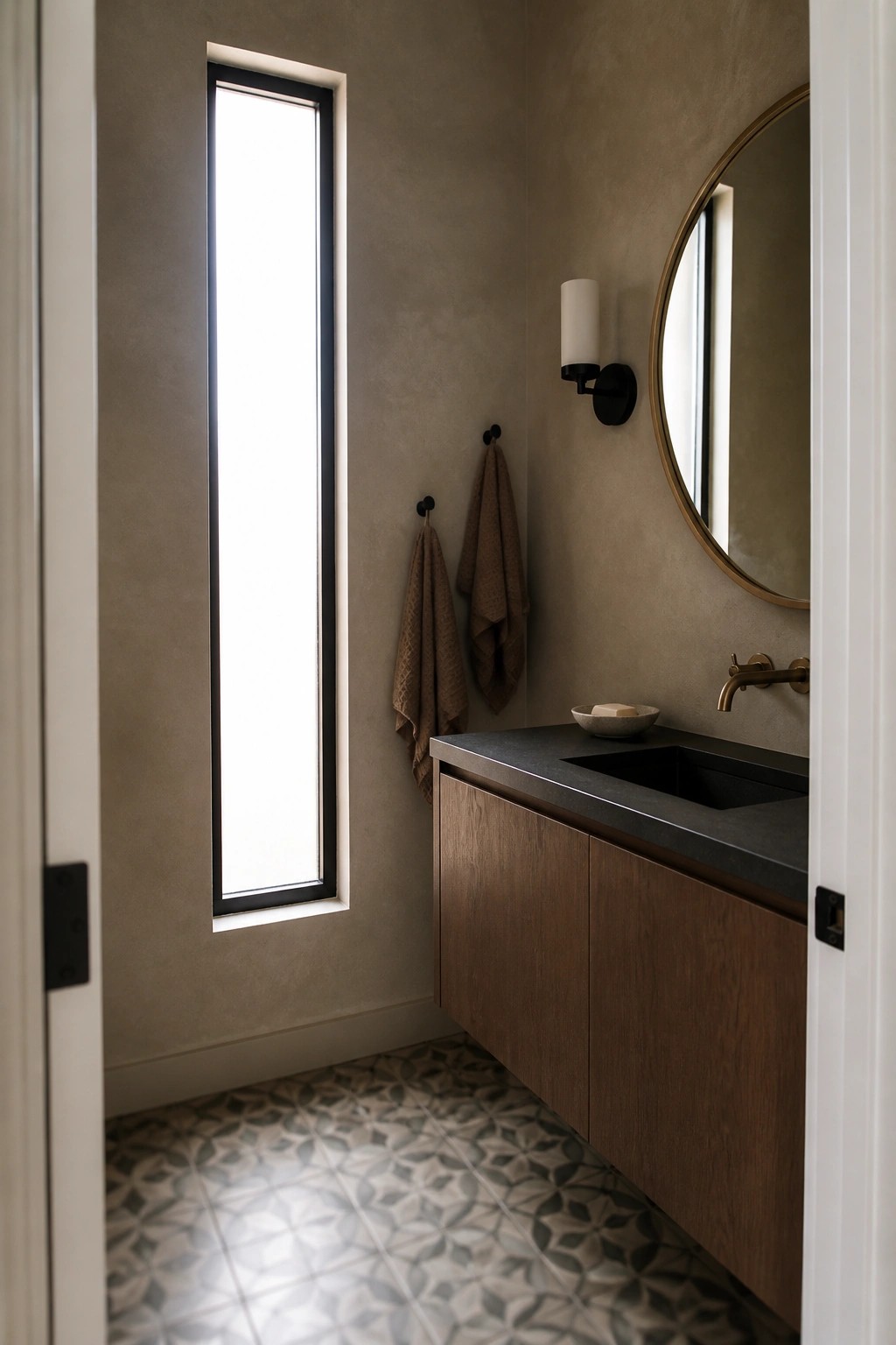 A bathroom interior with taupe walls, wood vanity, black stone countertop and sink, round mirror, wall sconce, and a tall narrow window.