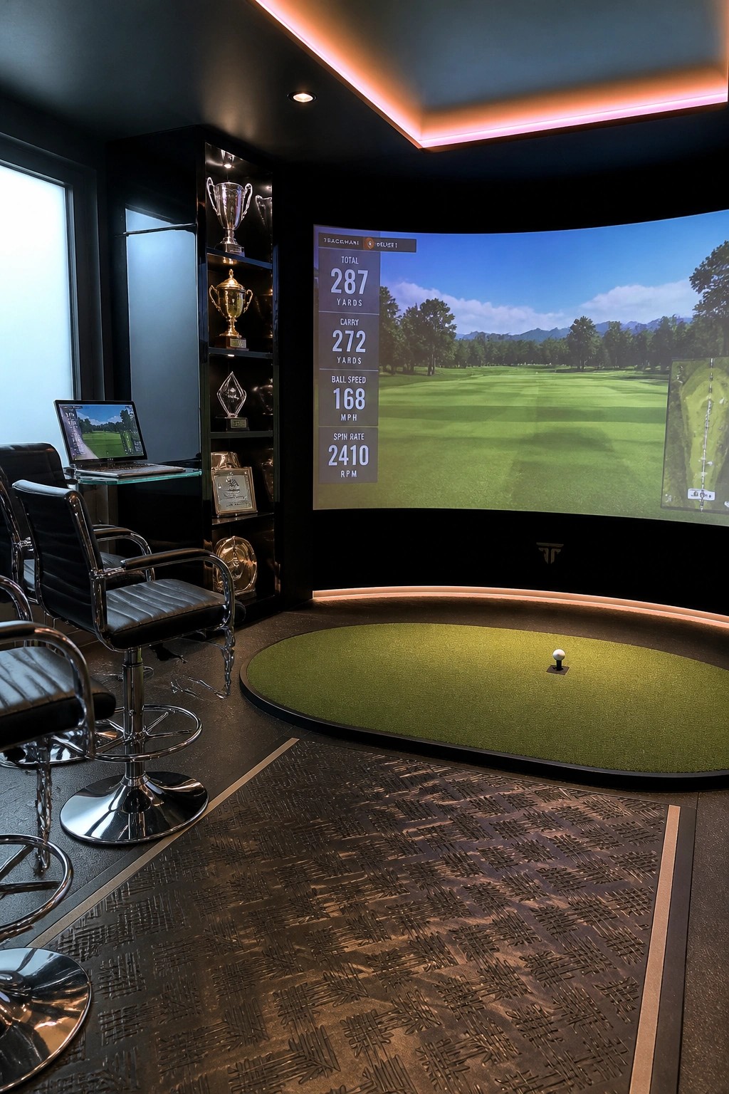 Indoor golf simulator room with a large curved impact screen showing golf stats and a course view, a small oval turf putting area with a ball, black bar stools, and a dark shelving unit holding trophies.