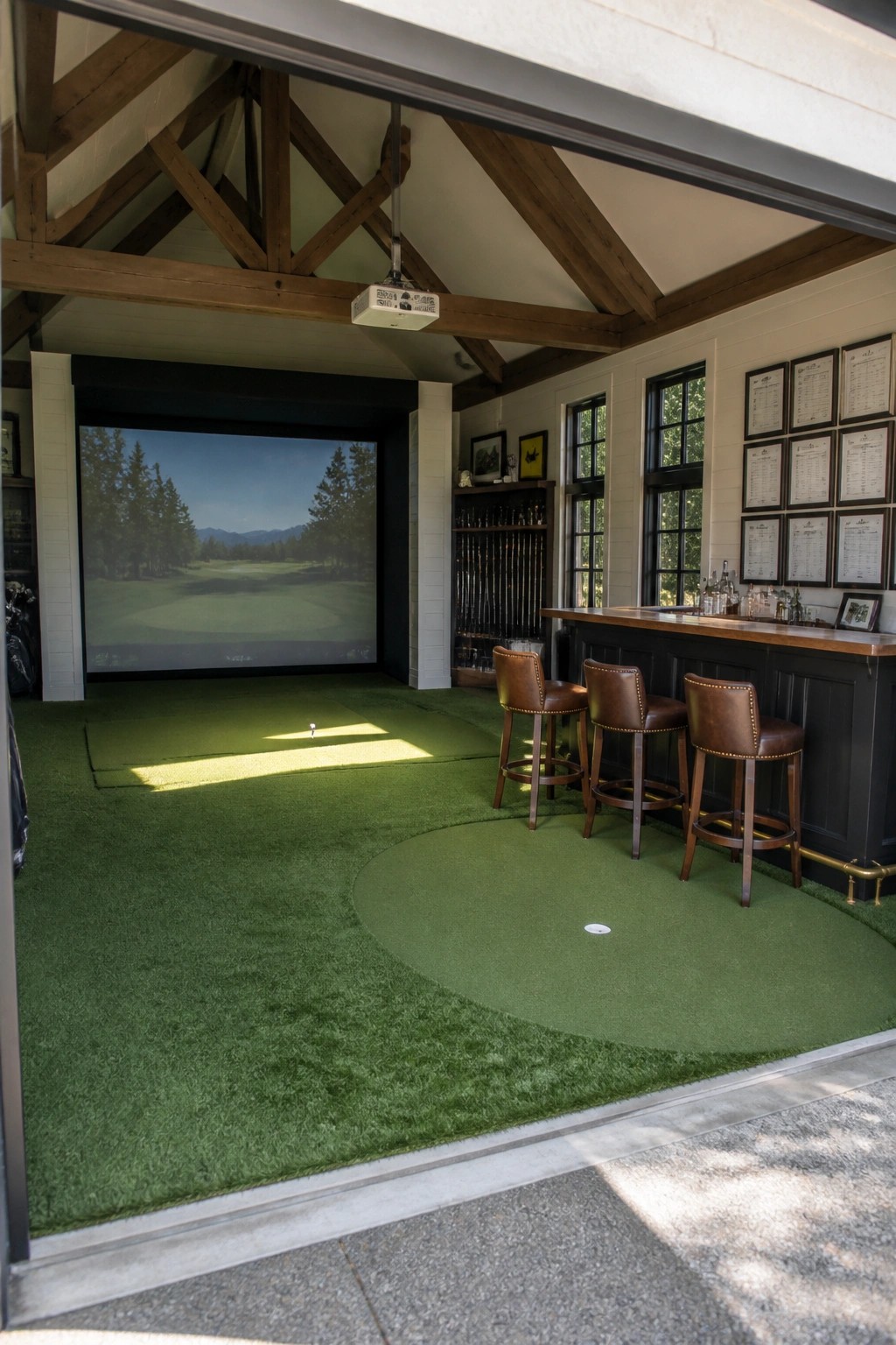 An indoor golf simulator room with a large impact screen showing a mountain golf course, full green artificial turf flooring, a dark wood bar with three brown leather stools, and exposed wooden ceiling beams.