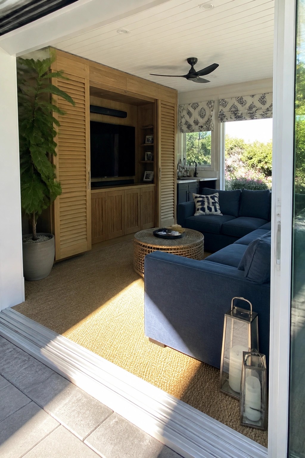 An interior view through open glass doors showing a blue sectional sofa, a round woven coffee table, and a wooden built-in media cabinet with louvered doors around a television.
