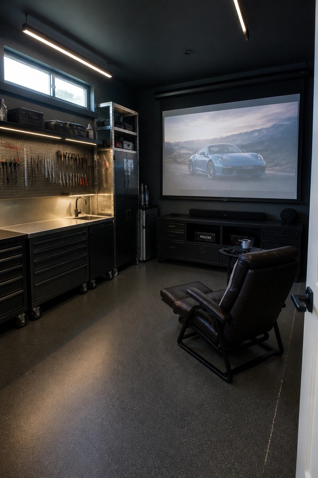 A dark modern man cave with black custom cabinets, a lit pegboard tool wall, stainless counter, large projection screen showing a blue Porsche, and a brown leather recliner on dark flooring.