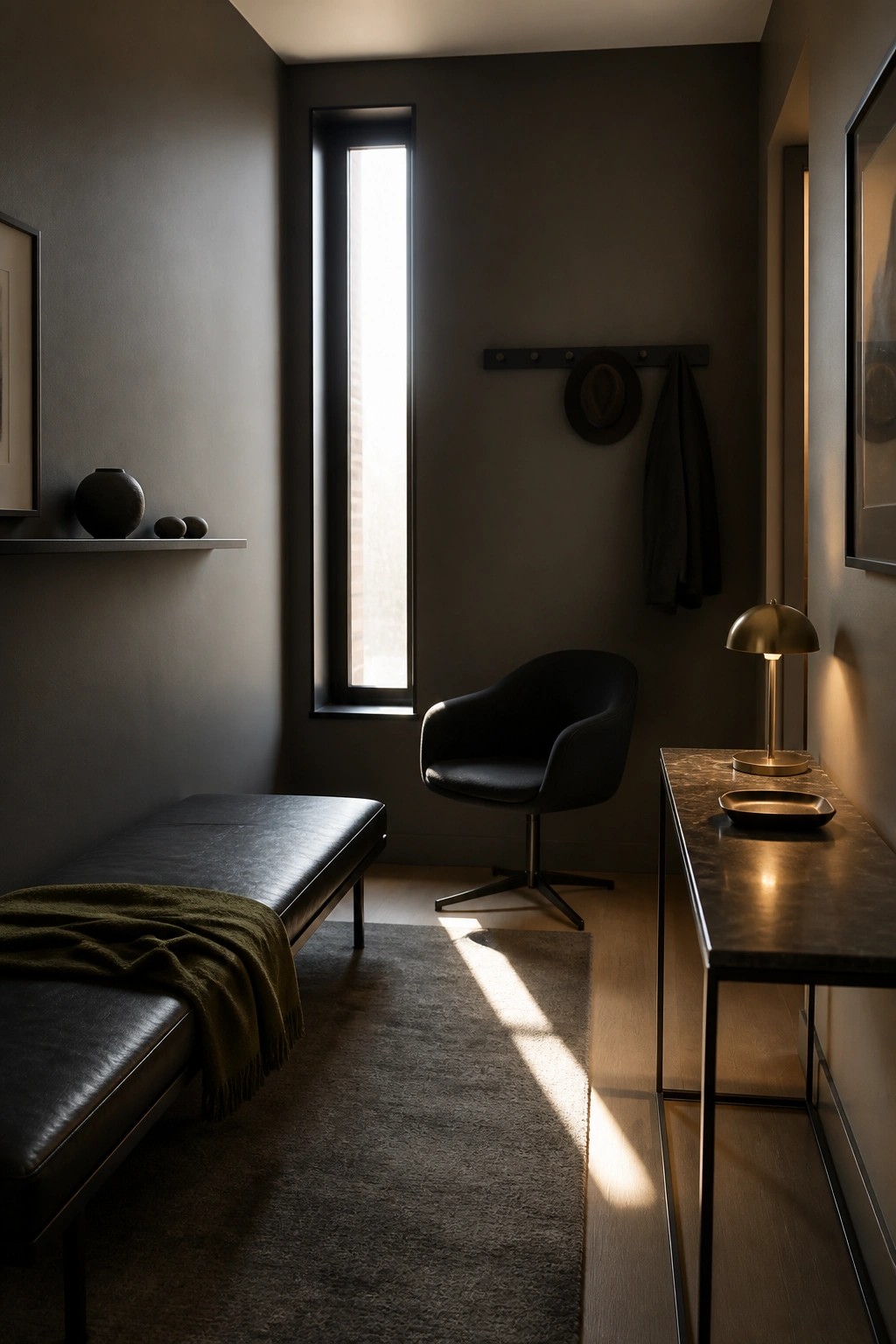 A narrow room with dark walls features a long black leather bench along the left wall, a black swivel chair near a tall window, and a dark marble console table on the right.