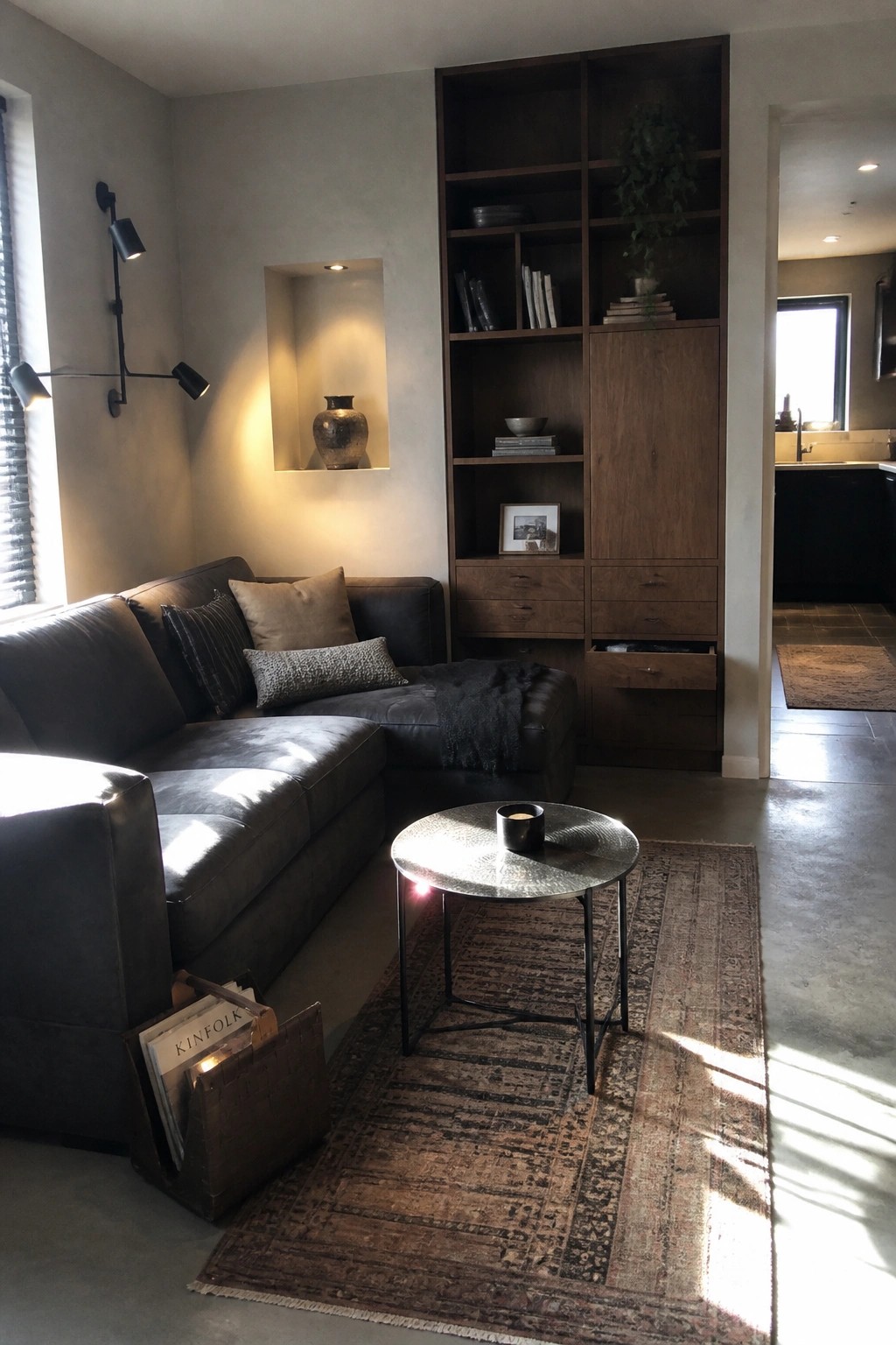 A small living room with a dark leather sectional sofa, a built-in dark wood shelving unit with cabinets and drawers, a round metal coffee table on a patterned rug, and a woven basket holding magazines.