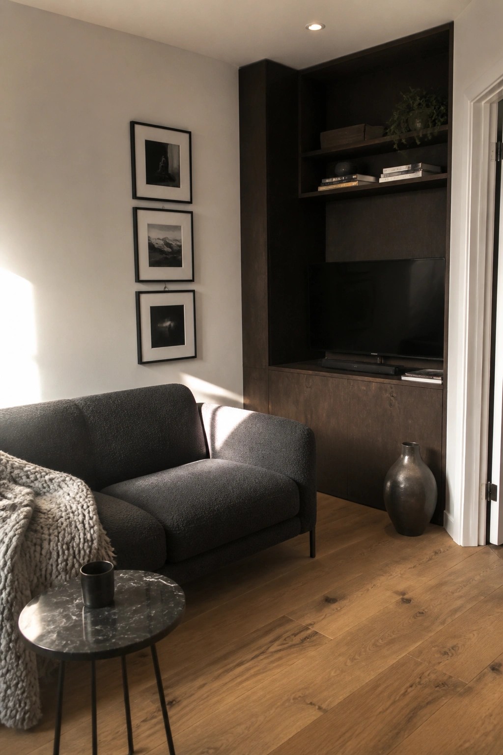 A small living room features a dark gray sofa, a built-in dark wood entertainment unit with television, and three framed photos on a light wall.