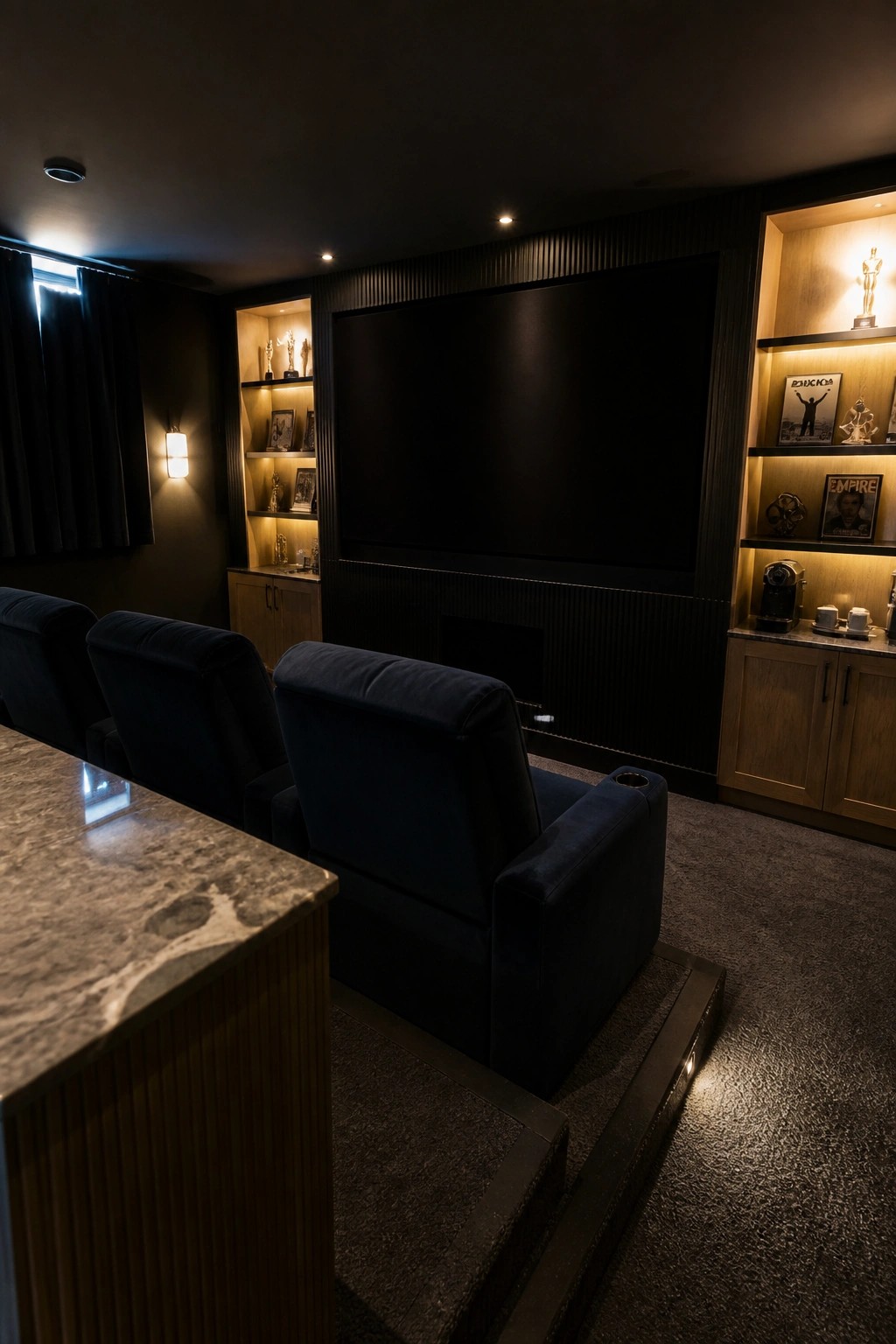 A basement home theater with three dark blue recliners on a raised platform facing a large screen, flanked by lit built-in shelves holding trophies and framed pictures.