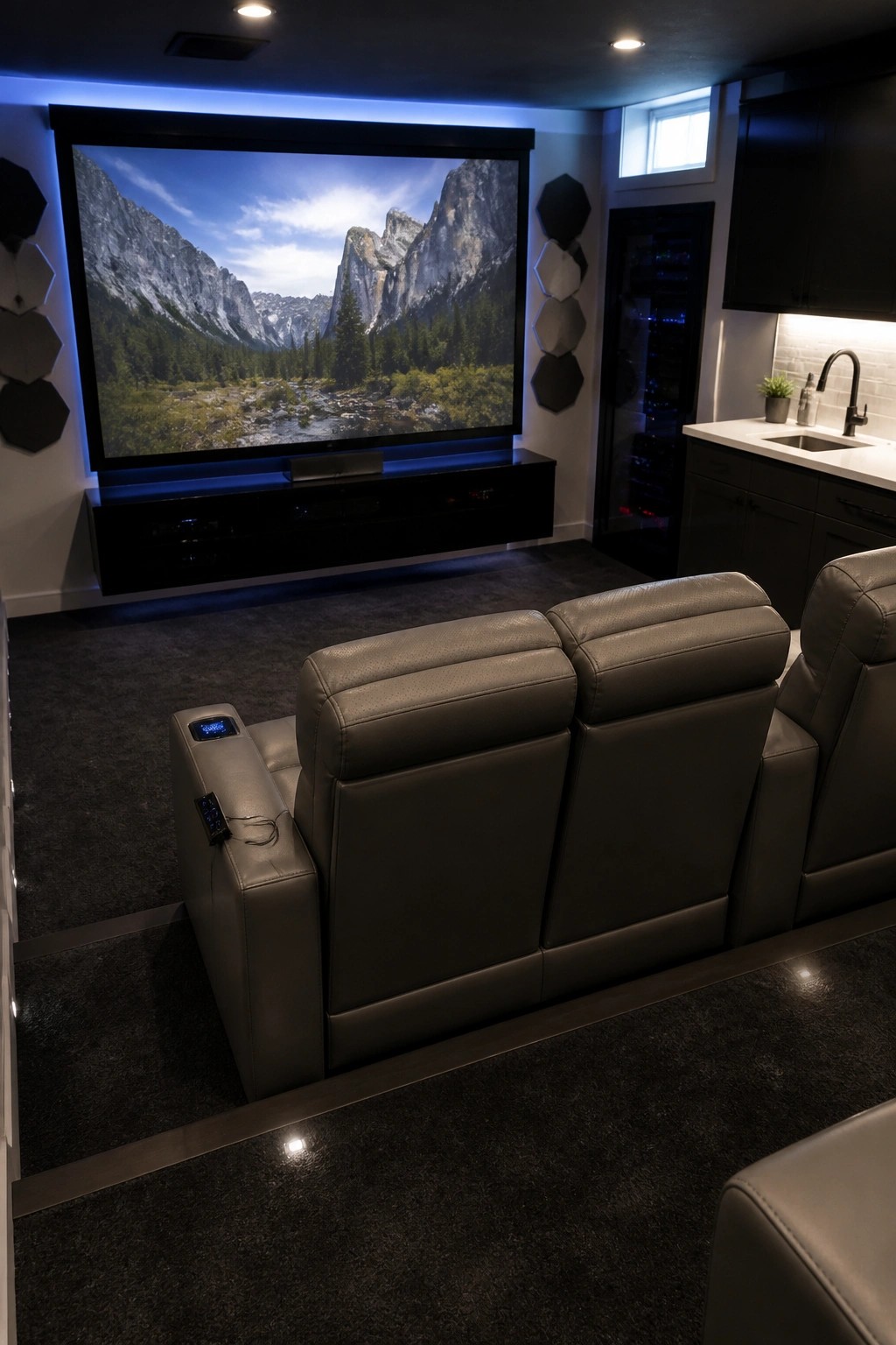 A basement home theater with gray leather reclining seats facing a large wall-mounted screen showing a mountain landscape, with dark carpet and a kitchenette area visible to the side.
