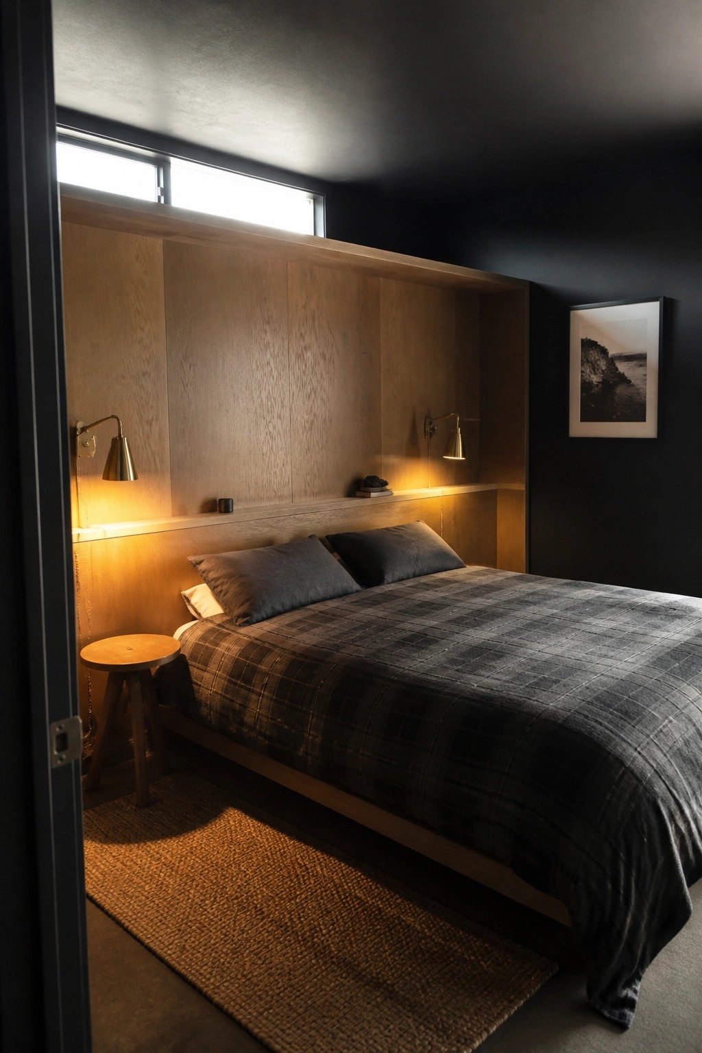 A bedroom featuring a bed recessed into a light wood alcove set against black walls, with two wall sconces and a high horizontal window above.