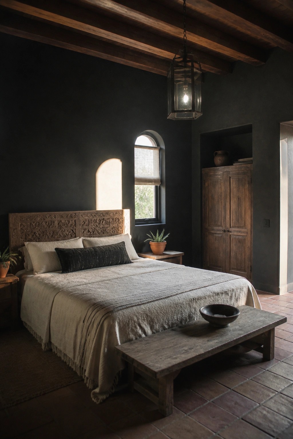 A bedroom with black walls features a large bed with a carved wooden headboard, a wooden wardrobe in a niche, terracotta tile floors, and a hanging lantern light fixture.