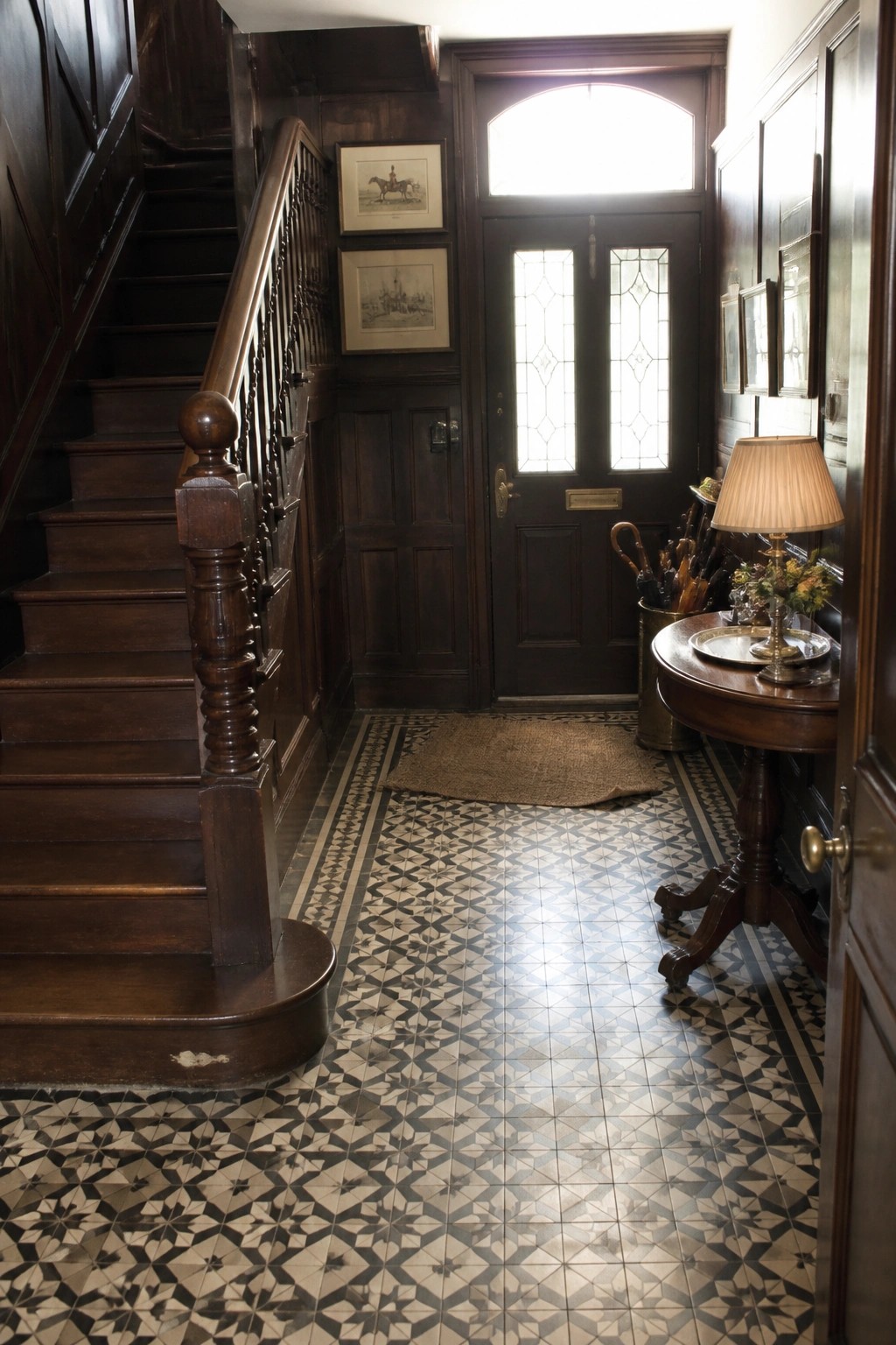 A traditional interior entry hall featuring a dark wood staircase, patterned tile floor, front door with glass panels, and a small wooden table with a lamp.