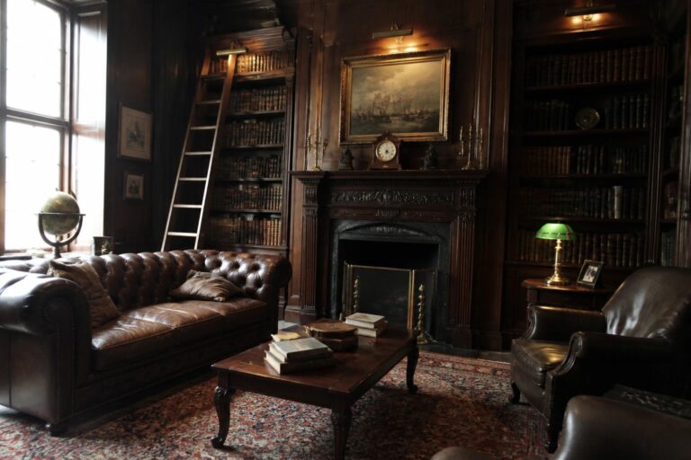 21 Classic Old Money Masculine Interiors Using Dark Wood and Traditional Furniture A classic study featuring dark wood built-in bookshelves, a brown leather sofa, a wooden coffee table with books, and a fireplace with a painting above it.