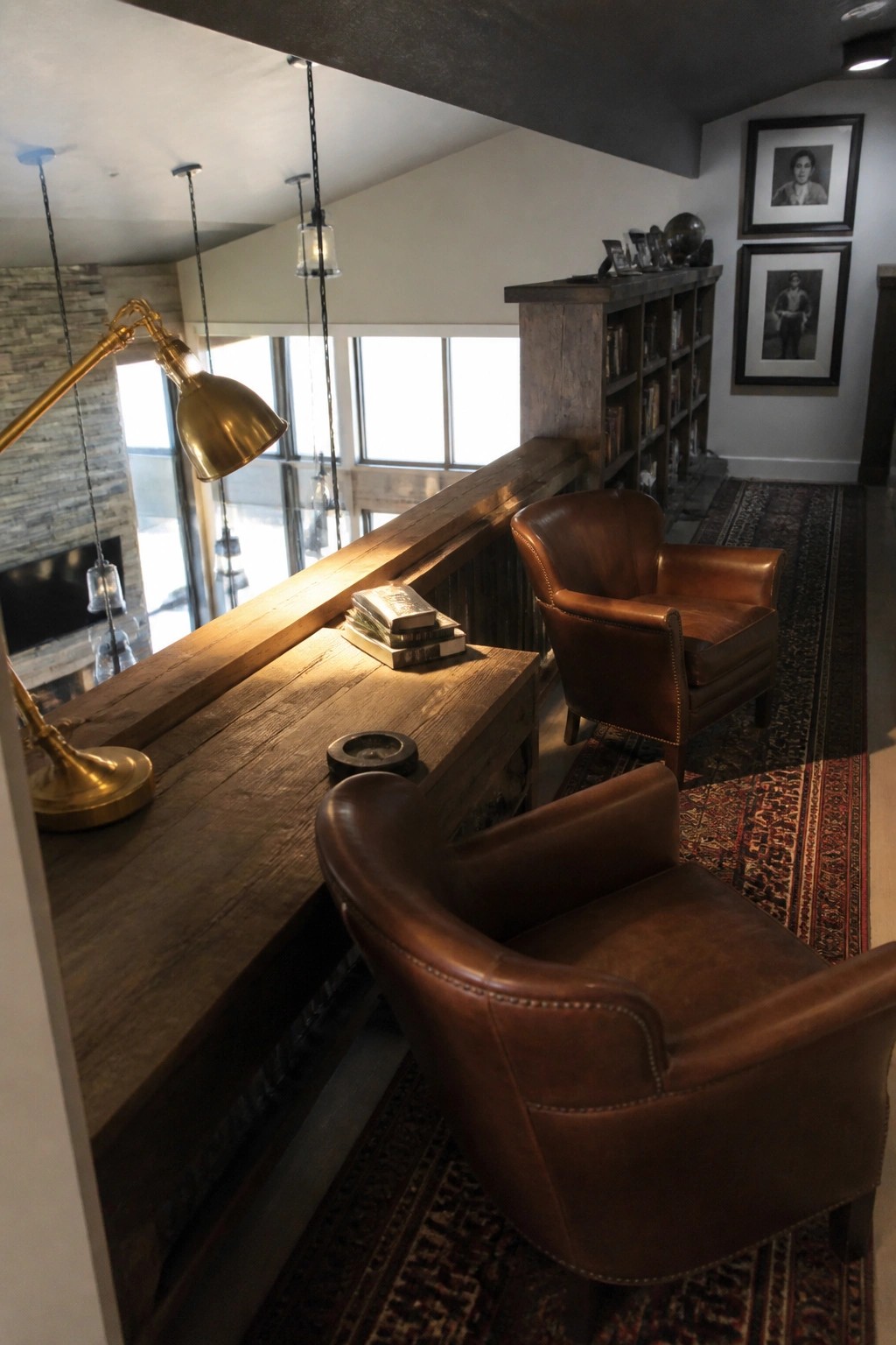Leather armchairs sit beside a long reclaimed wood desk or railing in a loft interior, with bookshelves and a fireplace visible in the background.