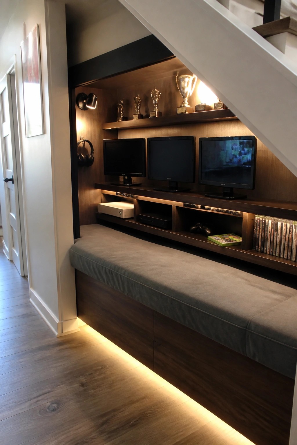 Recessed gaming area under stairs with three monitors above a long gray upholstered bench and open wooden storage shelves holding consoles and game cases.