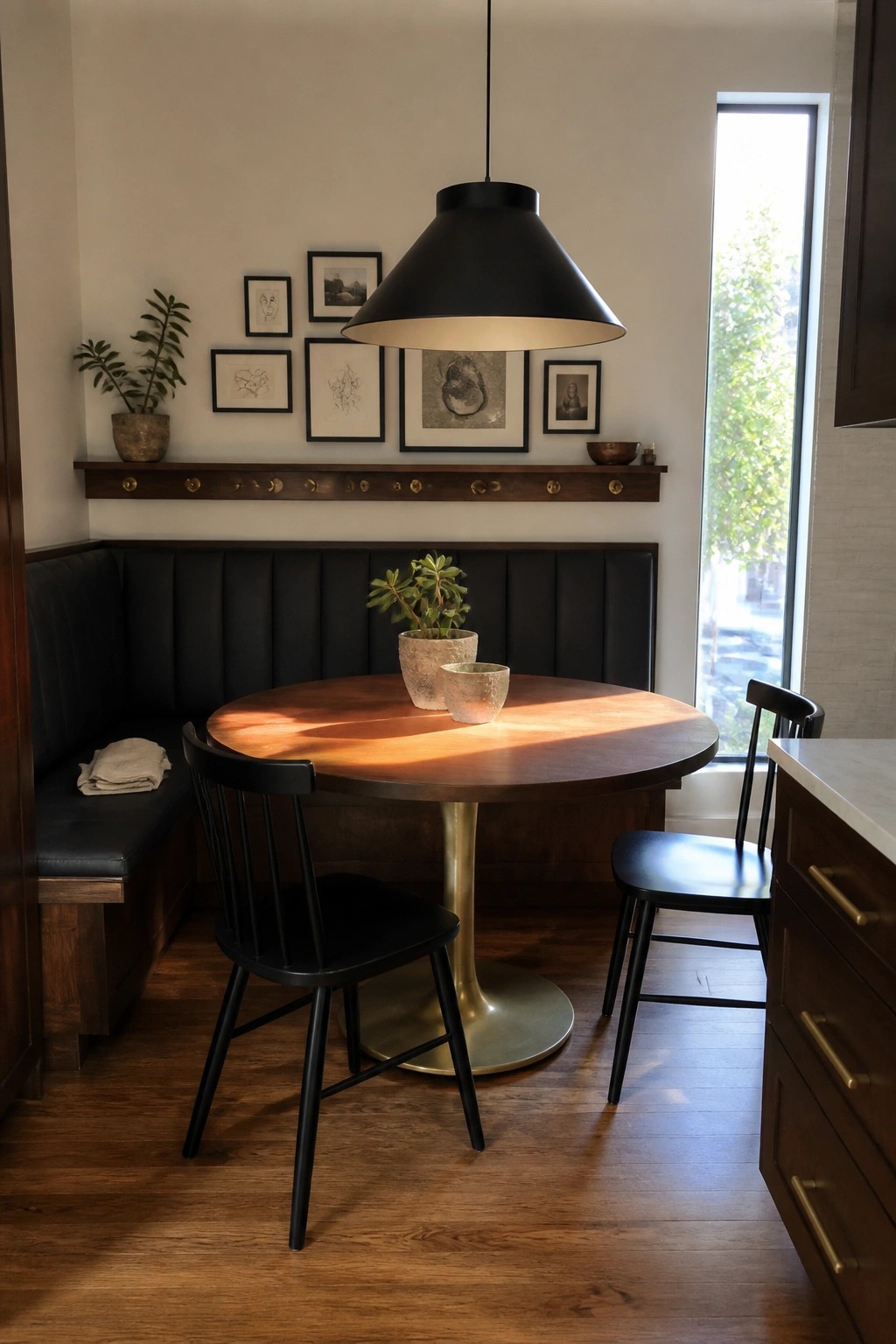 A round walnut dining table with a brass pedestal base sits in a corner with a black upholstered banquette, two black spindle chairs, and a large black pendant light overhead.