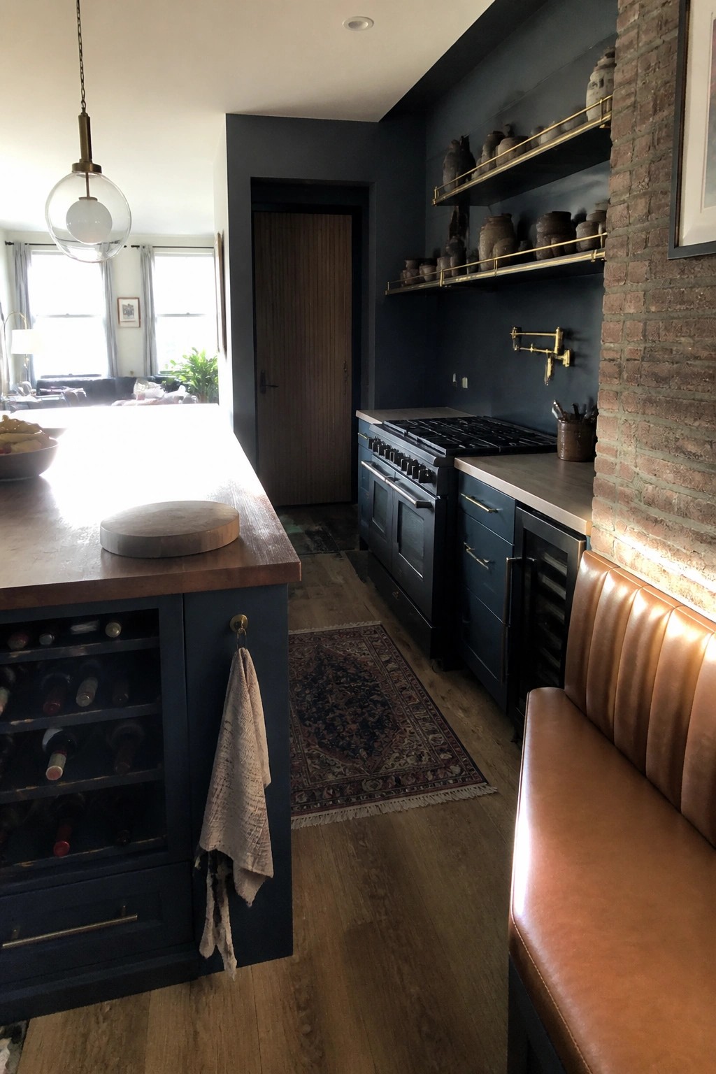 A kitchen interior with dark blue cabinets, a wooden island holding a cutting board, a gas range, brass open shelves, and a brown leather bench along a brick wall.