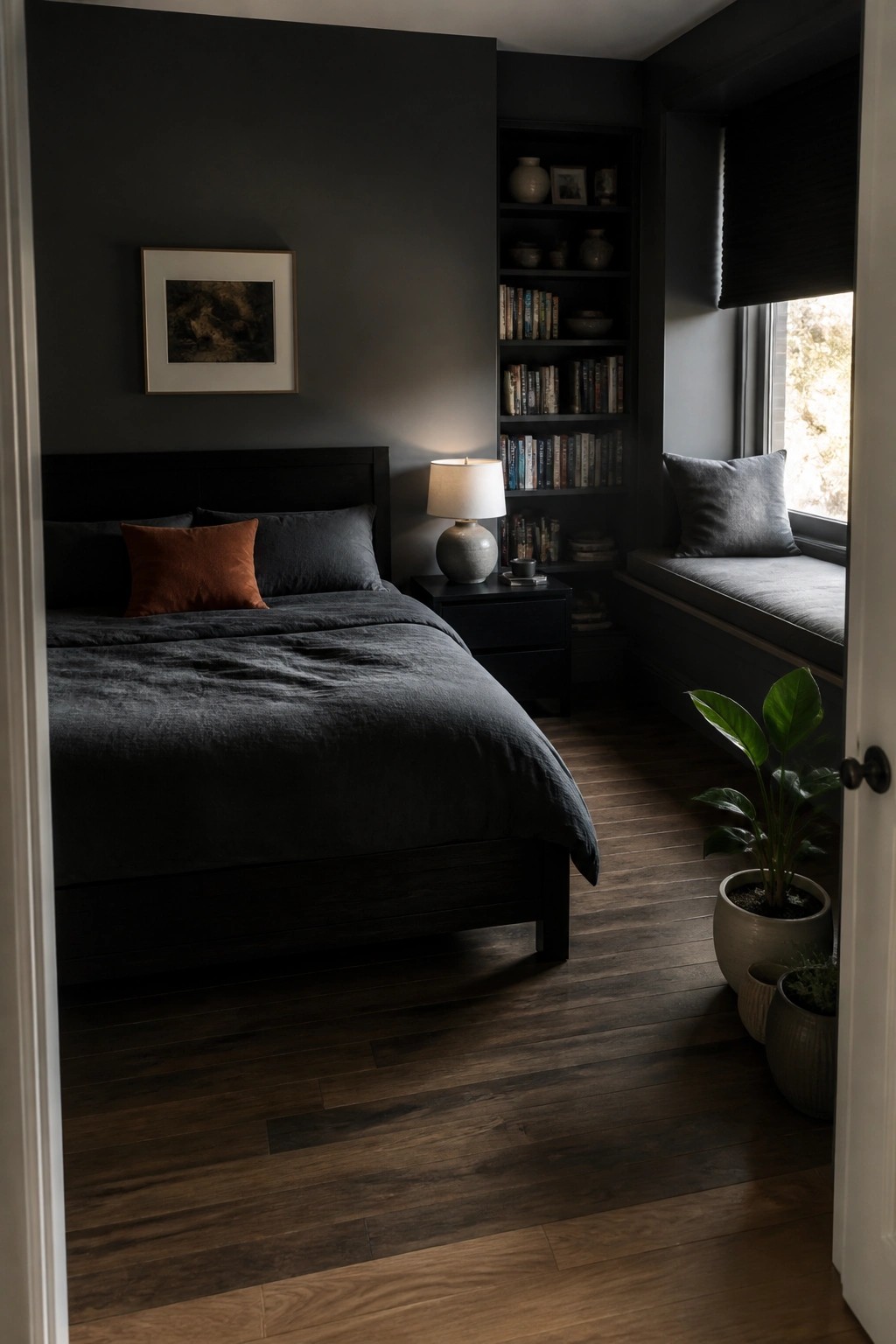 A dark bedroom with a bed covered in charcoal bedding, built-in bookshelves filled with books and small objects, a bedside lamp, and a window seat with a gray cushion.