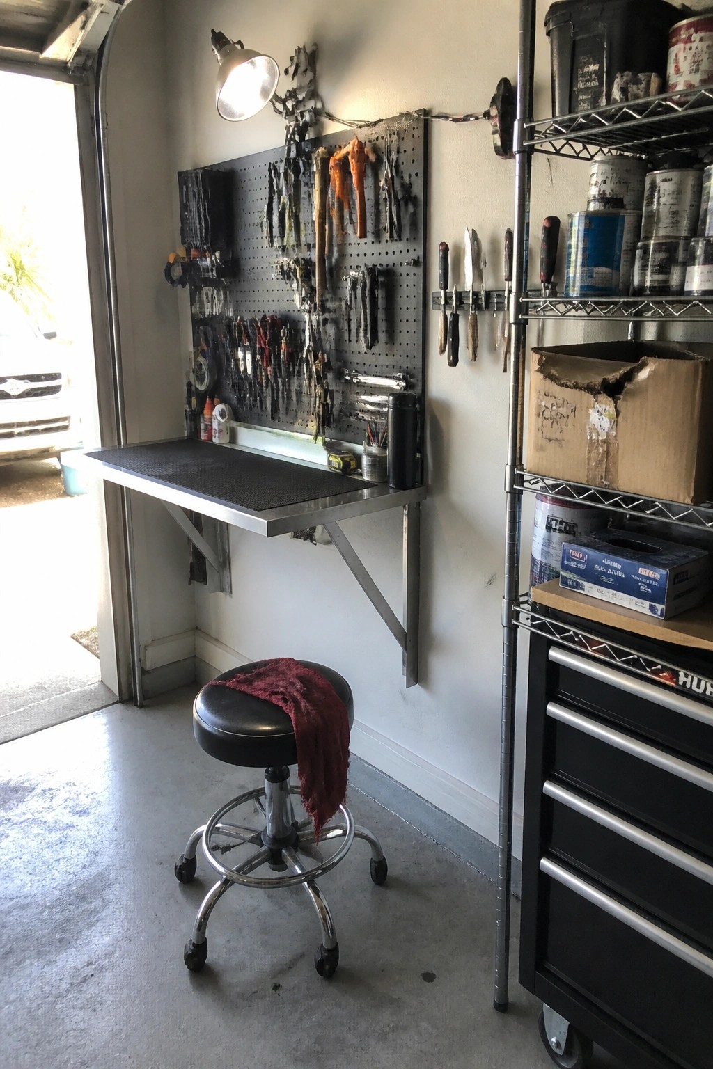 A compact garage workspace with a metal fold-down table, pegboard holding tools on the wall, a black rolling stool, and metal shelving with cans and boxes.