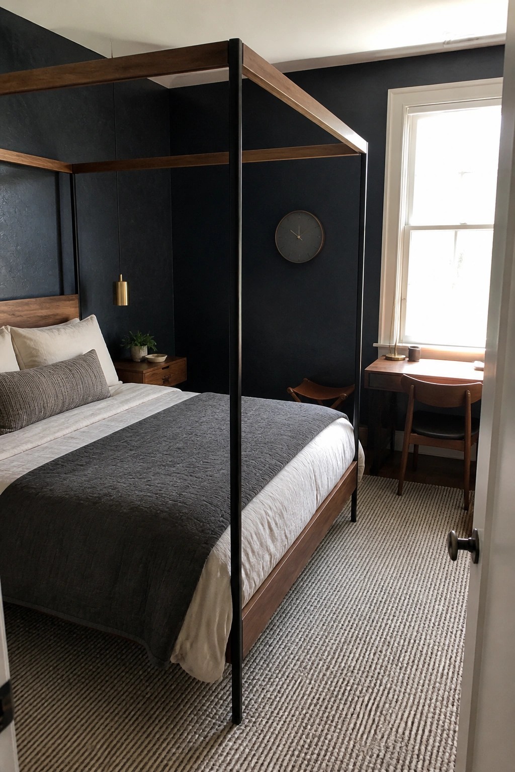 A bedroom with dark blue walls, a wooden four-poster bed with white and gray bedding, a wooden desk and chair near the window, and a small nightstand.