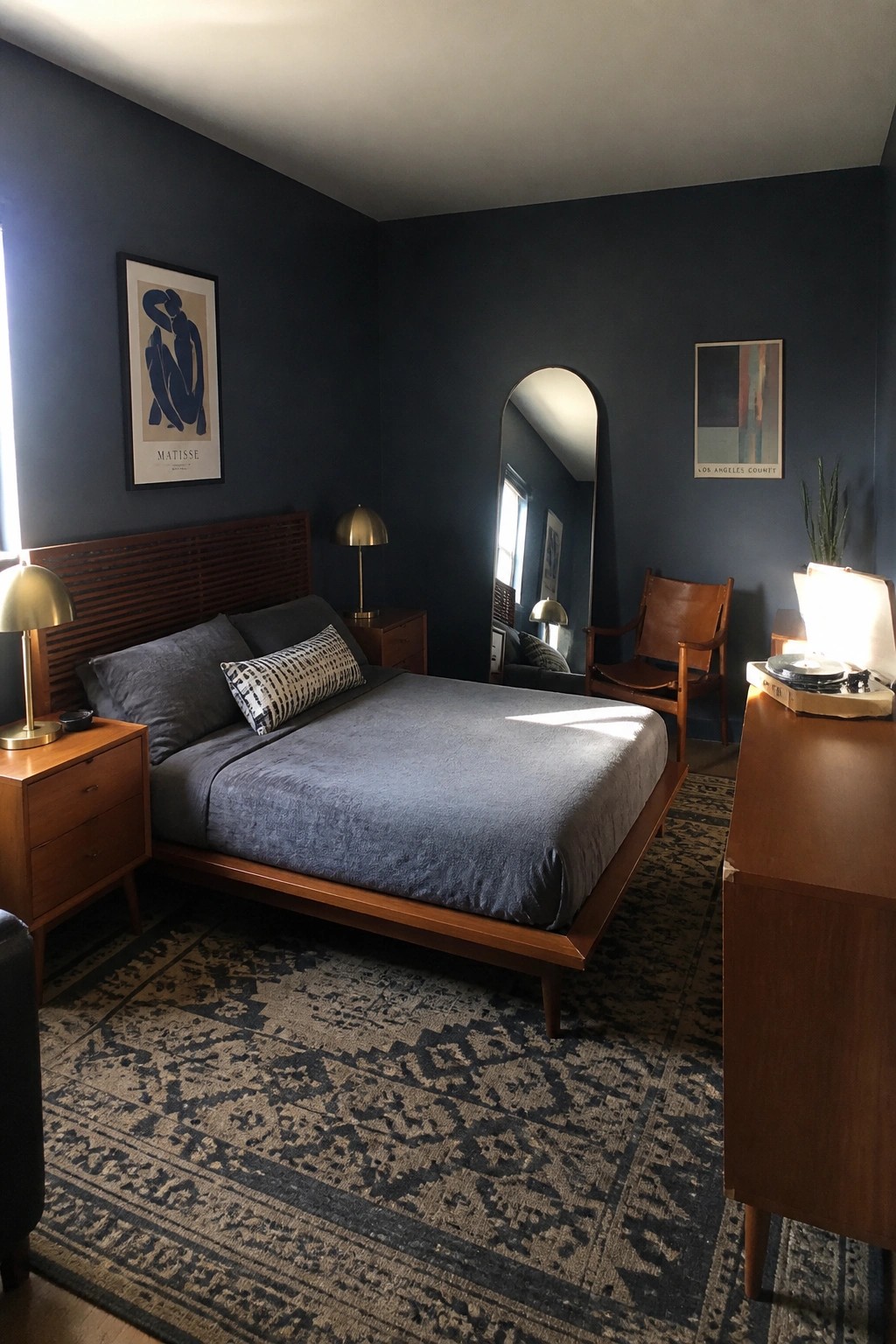 A bedroom with dark blue walls, a wooden bed with gray bedding, two wooden nightstands with brass lamps, a tall arched mirror, a wooden dresser holding a record player, and a patterned rug on the floor.