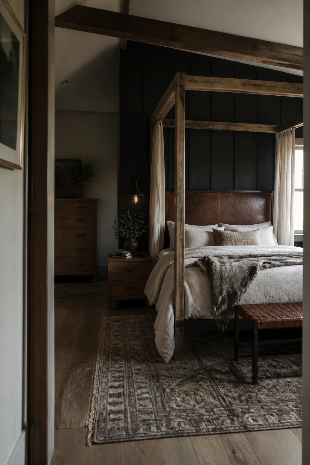 A bedroom featuring a four-poster bed with a brown leather headboard against dark paneled walls, layered neutral bedding, a wooden nightstand, and a patterned rug on hardwood floors.